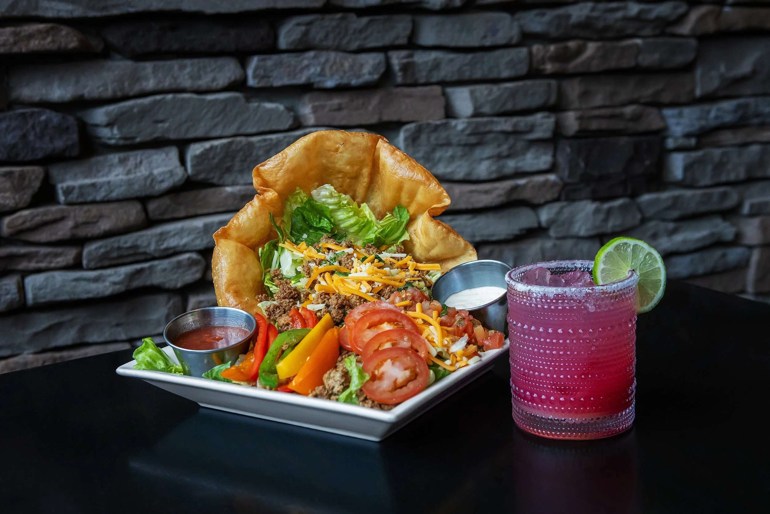Plate of taco salad with lettuce, tomatoes, shredded cheese, ground beef in a crispy tortilla bowl, and assorted vegetables, with two small containers of sauce, and a pink cocktail garnished with a lime slice.