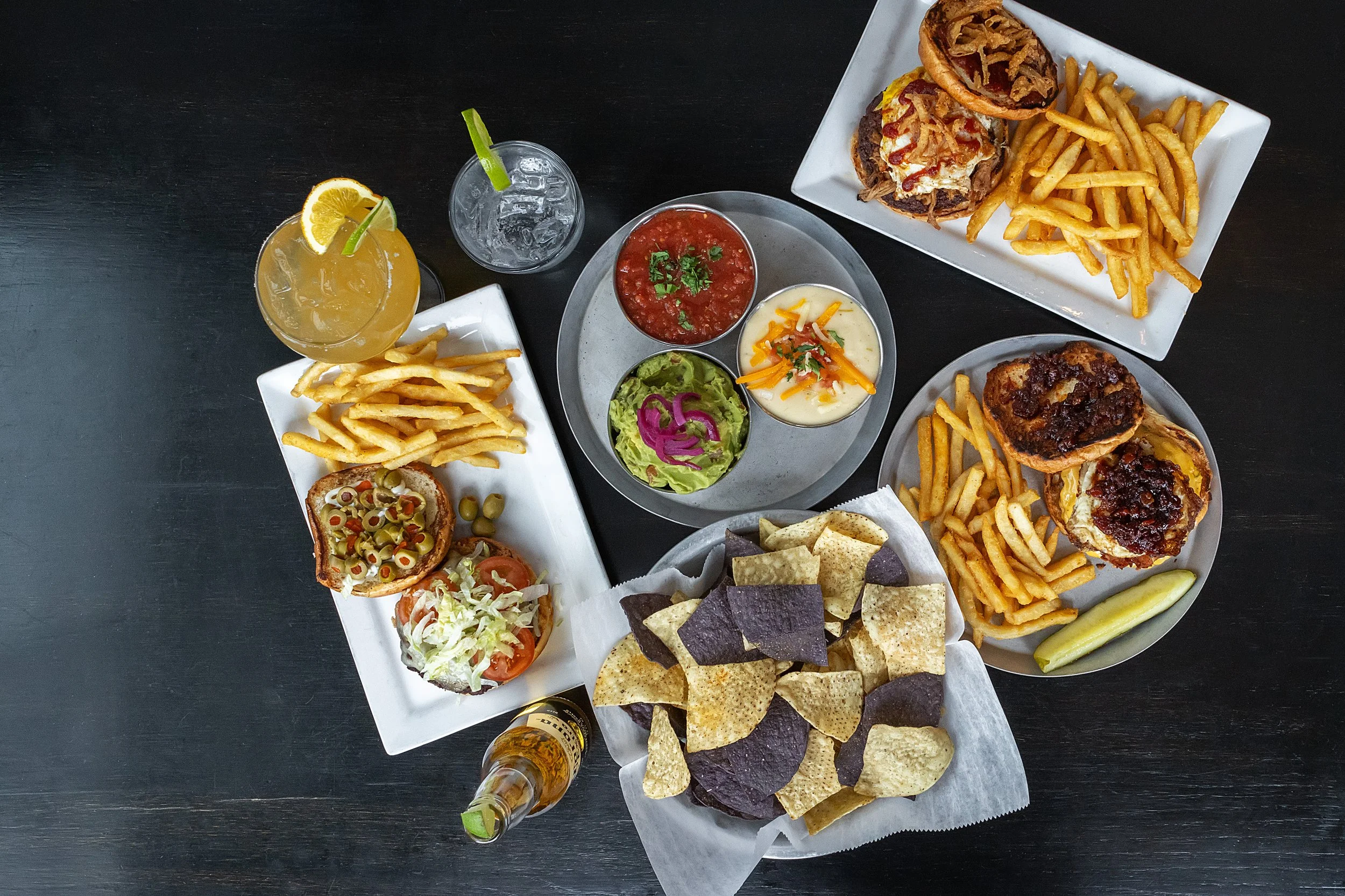 A table with Mexican food including sliders topped with olives, lettuce, and tomato, French fries, chips with salsa, guacamole, and sour cream, beer, and mixed drinks, alongside bowls of salsa, guacamole, and sour cream with shredded cheese, and quesadillas with beef and cheese.