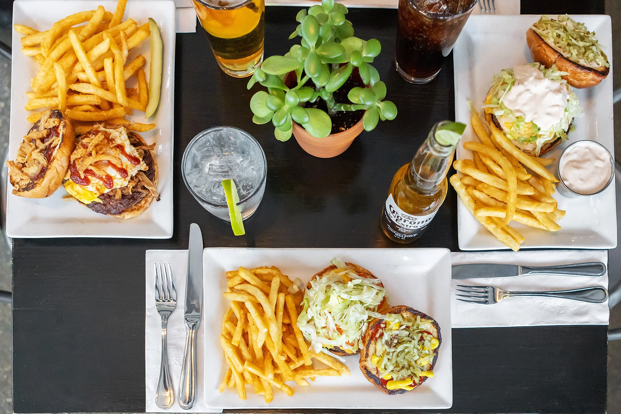 Three rectangular white plates with burgers, French fries, and side sauces on a black table. The plate at the top left has a burger topped with lettuce, tomato, onions, ketchup, and French fries, with a pickle on the side. The plate at the top right 
