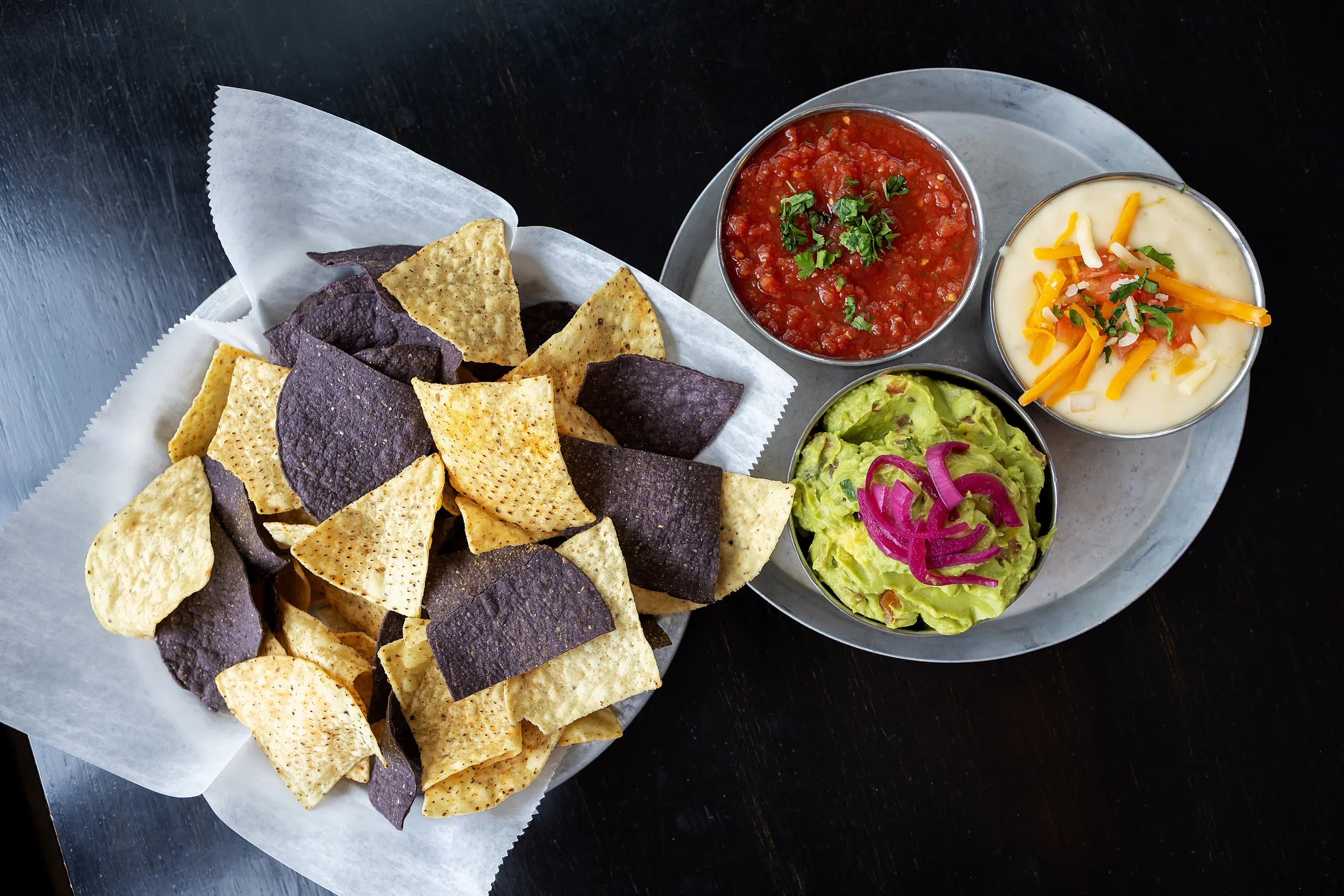 A basket of tri-color tortilla chips served with three small bowls of side dips: salsa, guacamole with pickled red onions, and queso cheese with shredded cheddar and cilantro, on a metal tray.