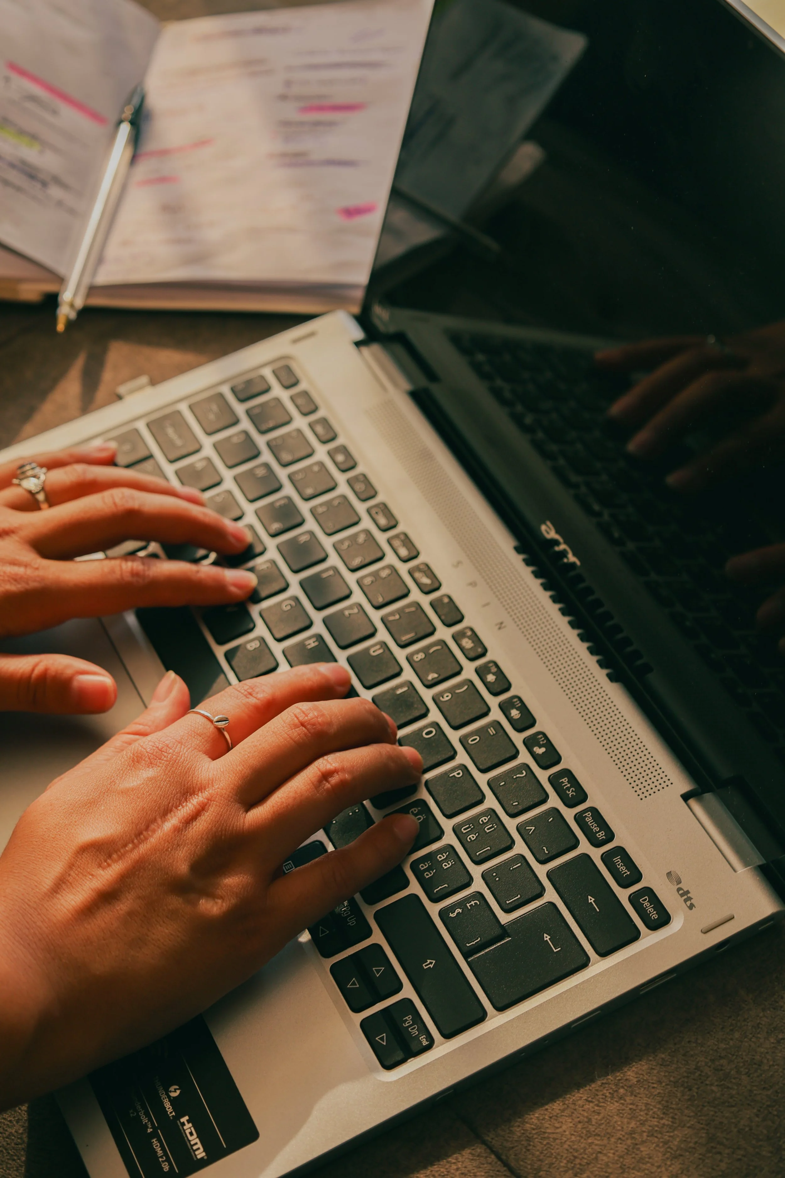 Hands typing on a laptop keyboard with a notebook and pen in the background.