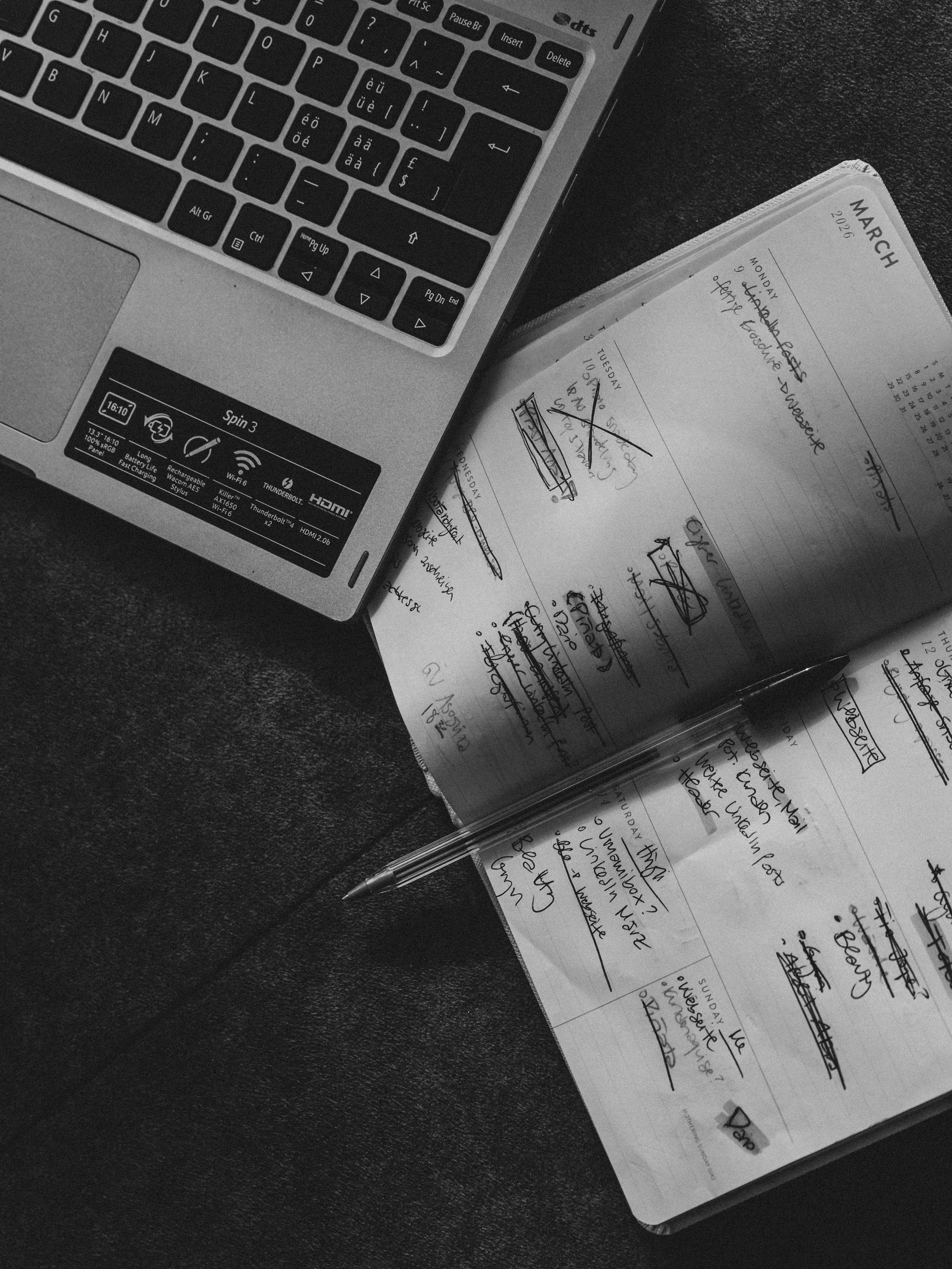 Black and white photo of an open planner with handwritten notes, a pen resting on the pages, and a laptop keyboard visible in the top left corner.