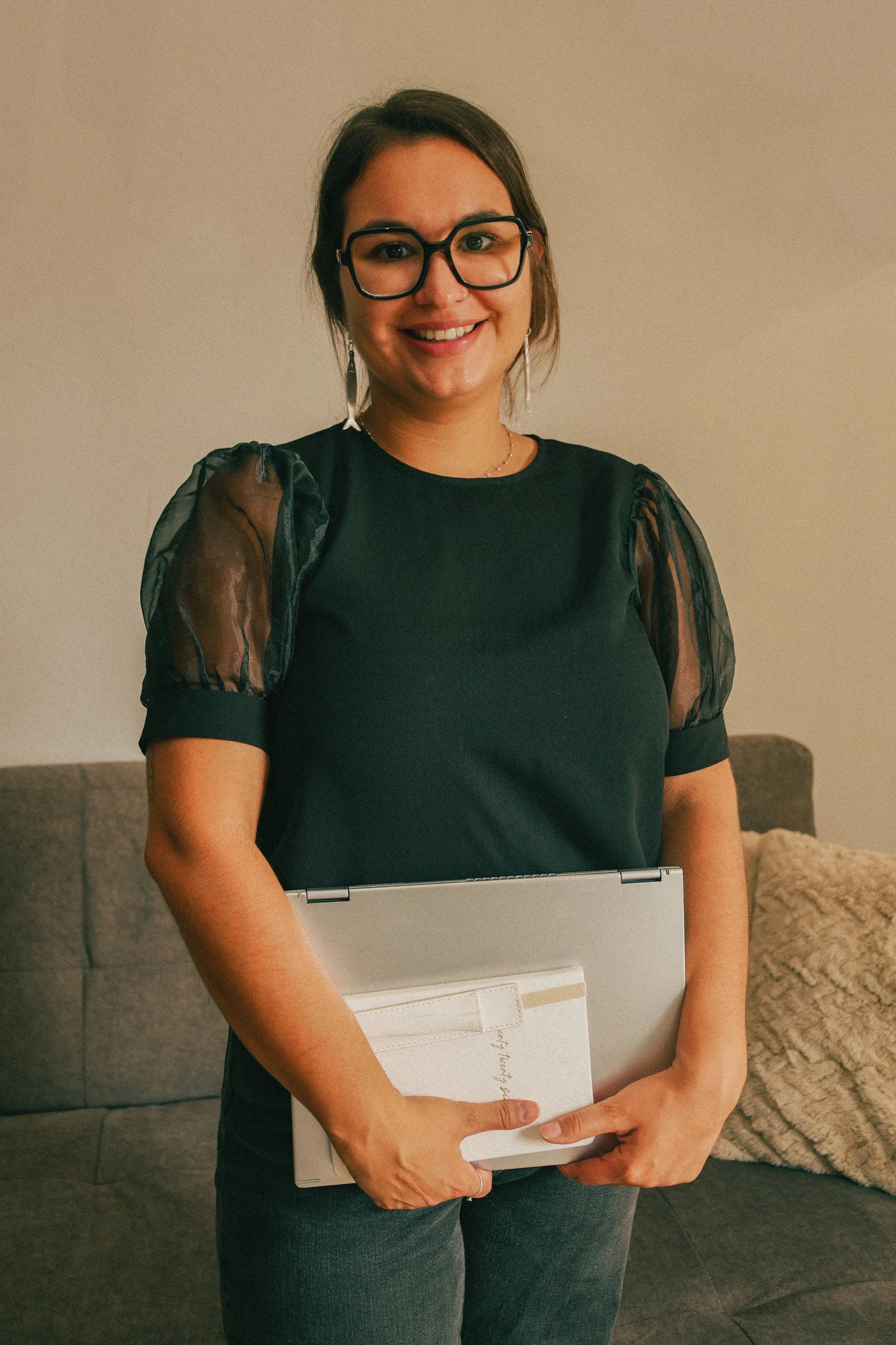 A business woman smiling while holding a closed silver laptop against her waist, standing in a living room with a brown couch and a textured pillow.
