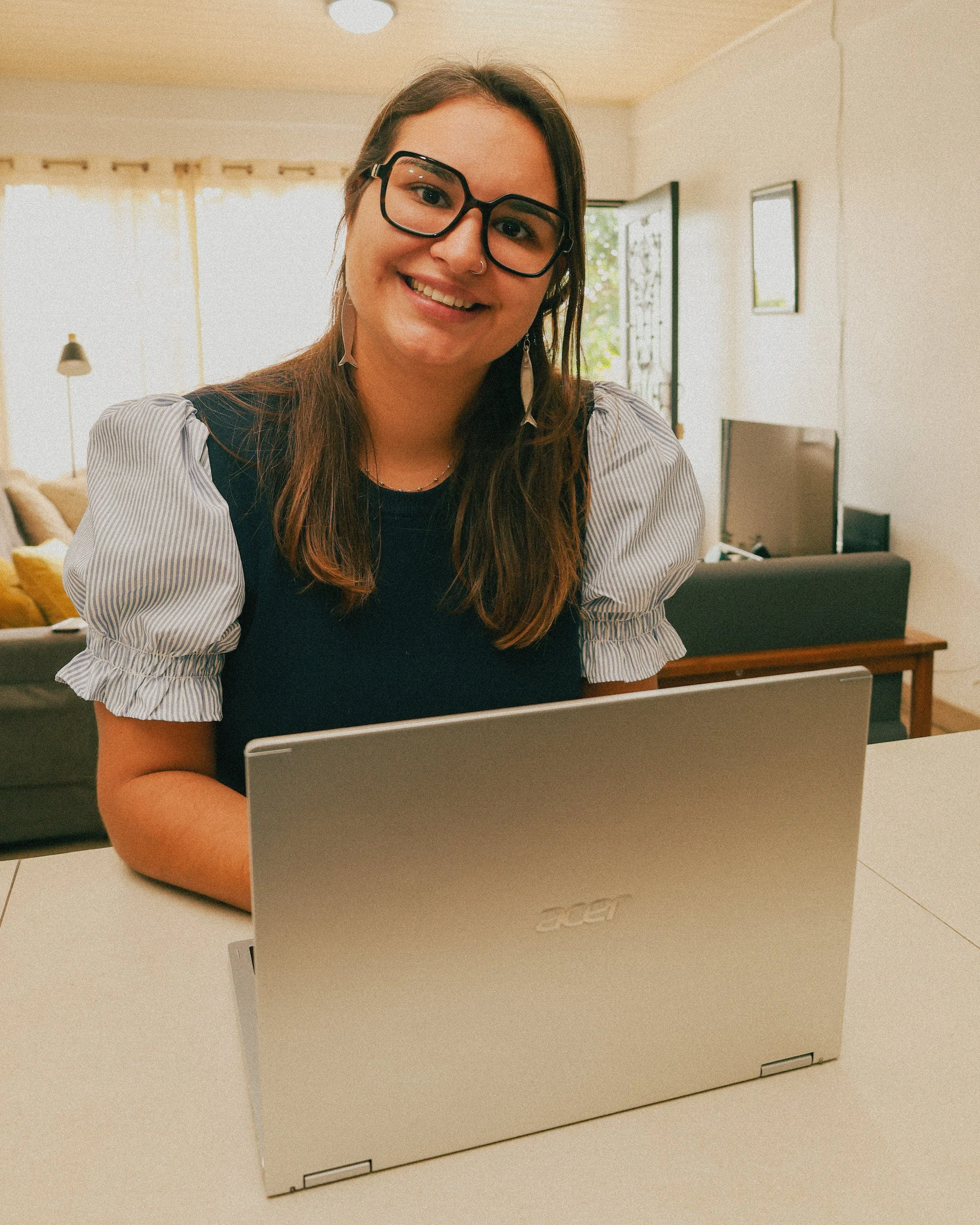 A young business woman with glasses and a nose ring sitting at a table with an open laptop, smiling in a well-lit living room.