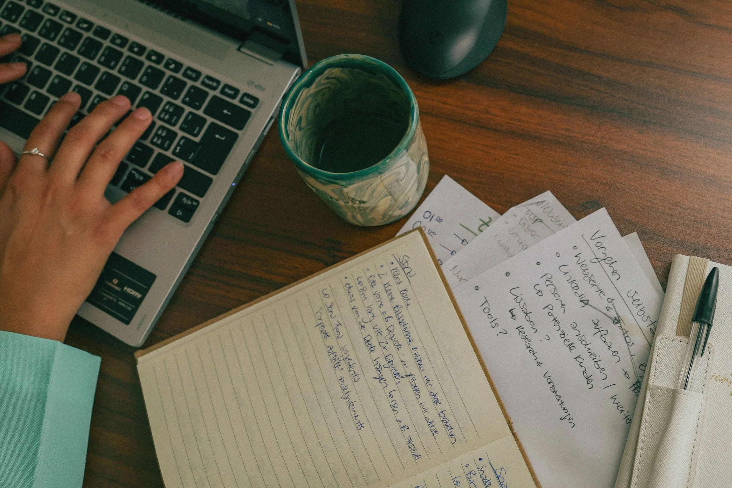 A person typing on a laptop with handwritten notes and papers scattered on a wooden desk, along with a colorful mug and a pen holder with a pen inside.