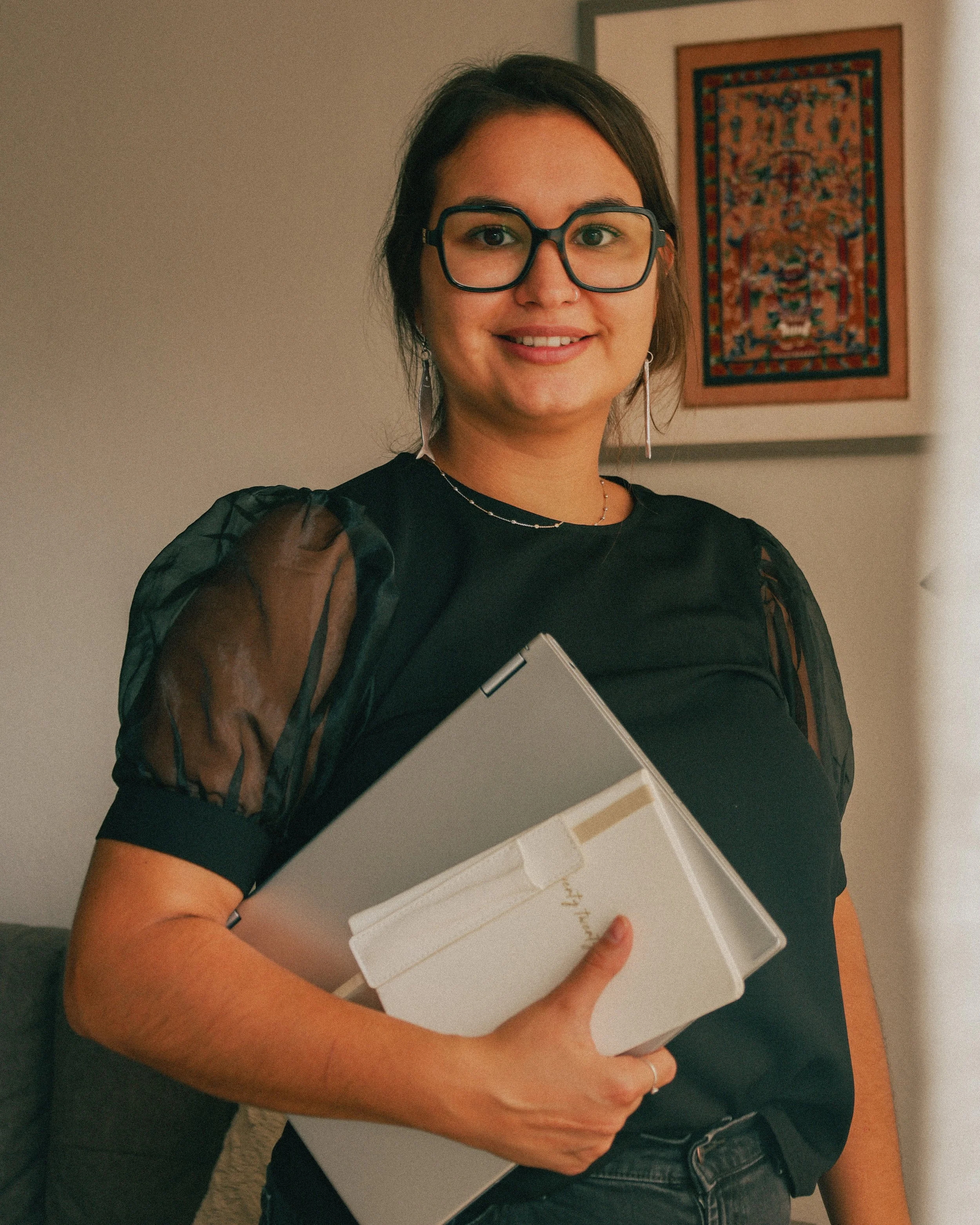 A woman with glasses holding a closed laptop and a white folder, standing indoors near a wall with framed artwork.