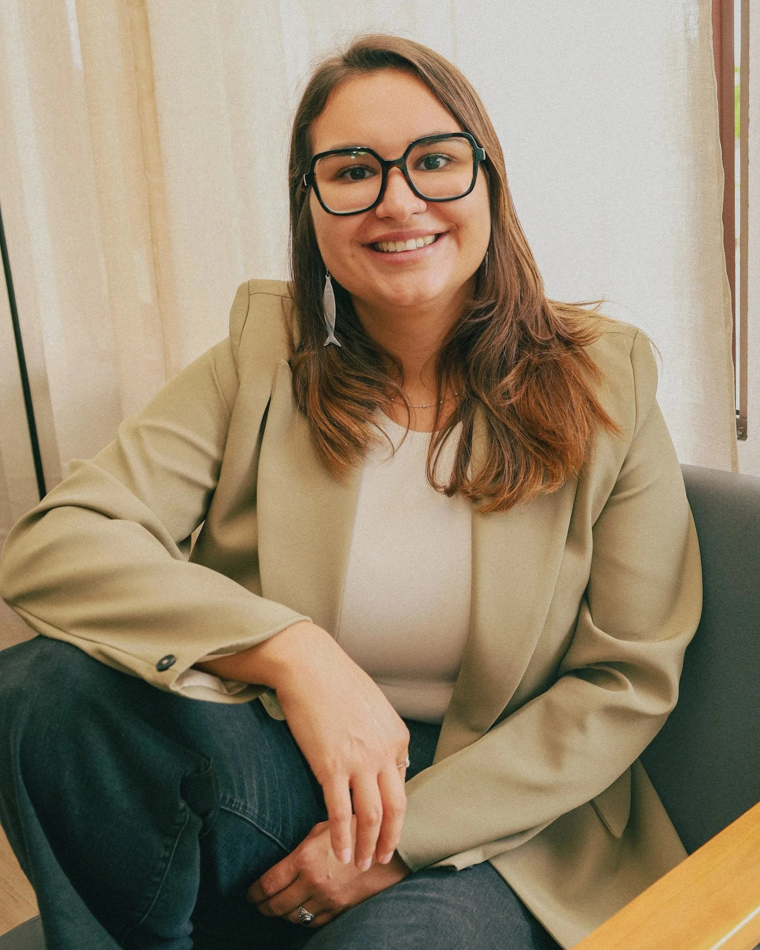 A woman with glasses, wearing a beige blazer and white top, sitting in a room with curtains, smiling at the camera.
