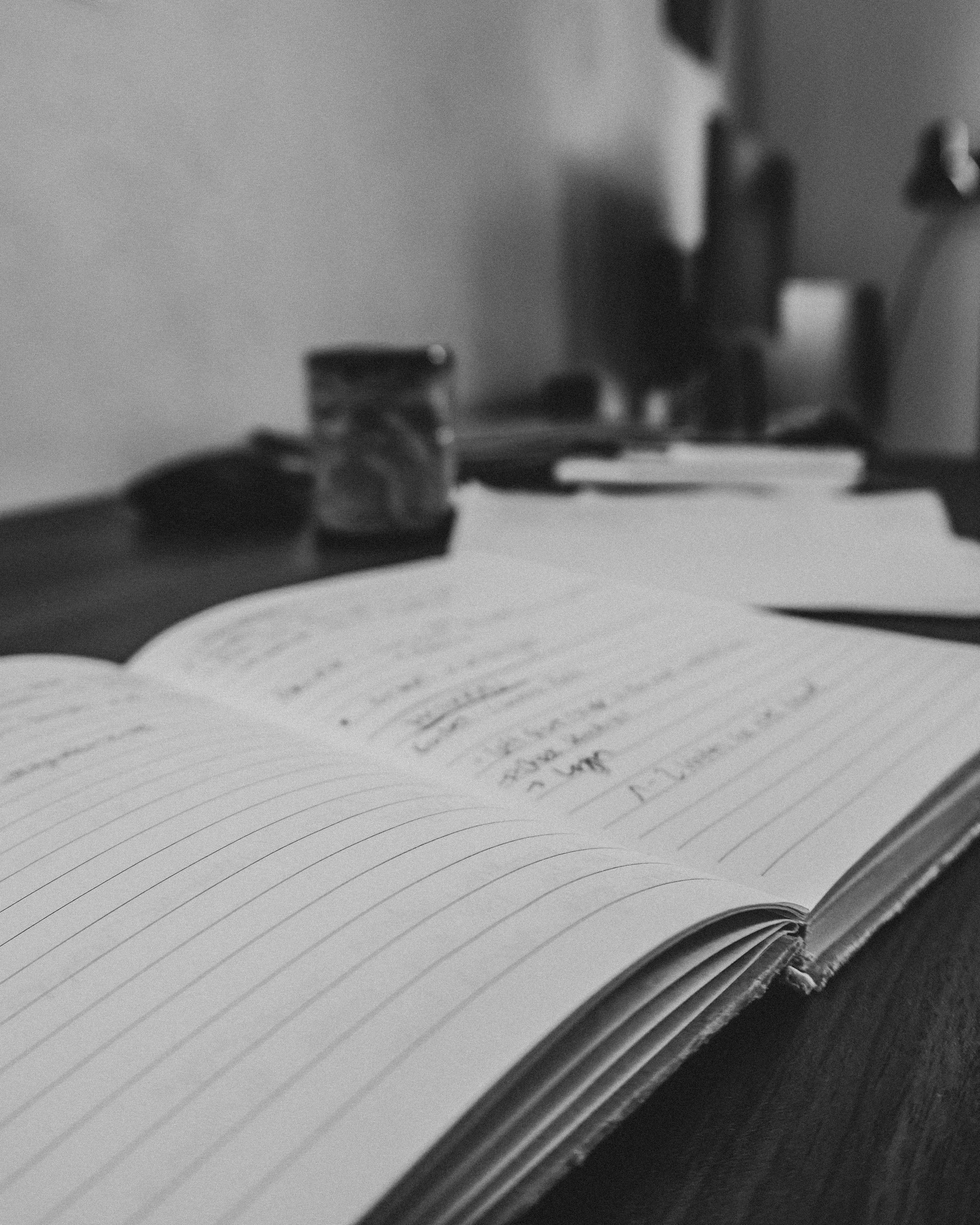 Open notebook with handwritten notes on a desk, with blurred objects including a jar, notebook, and a lamp in the background.