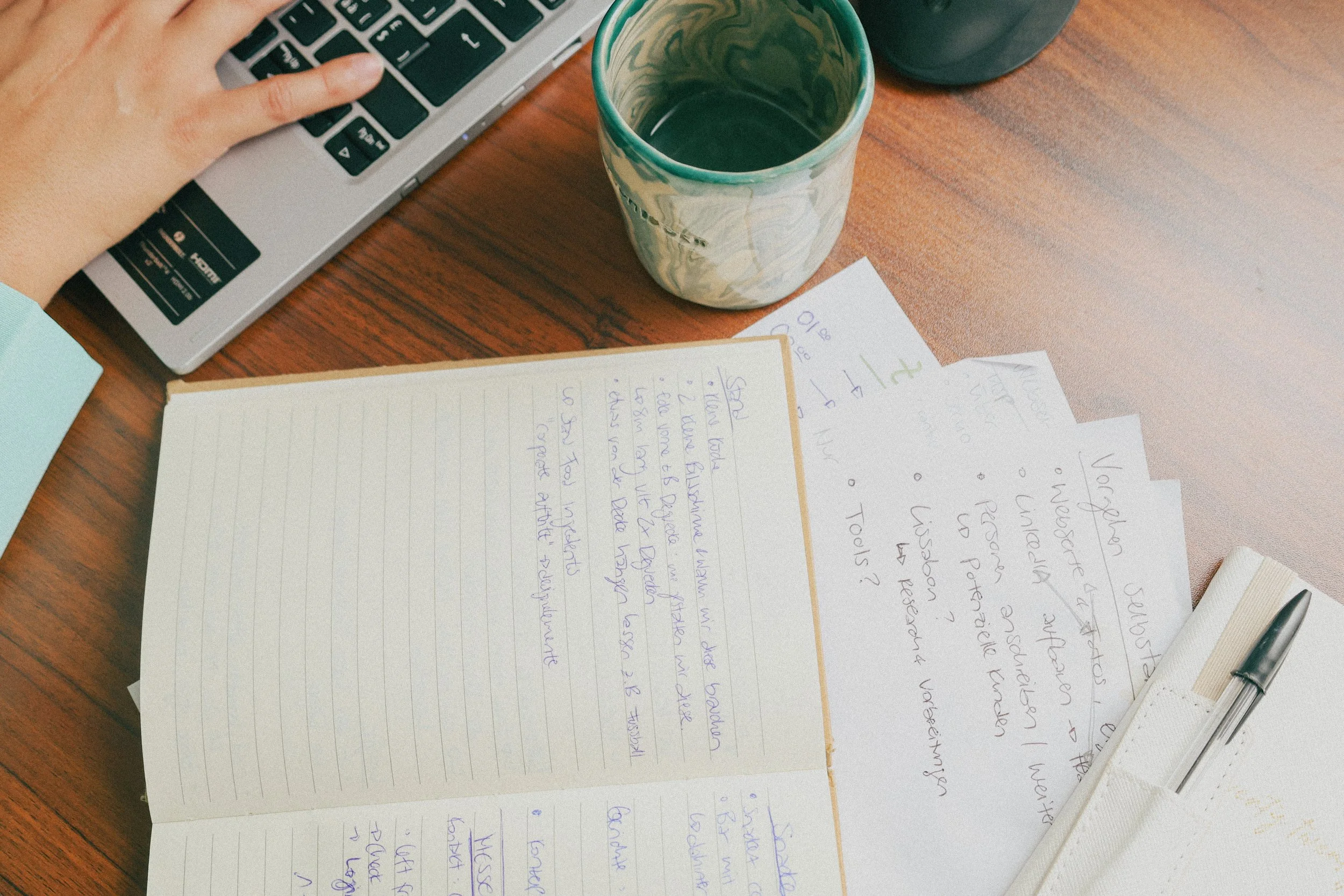 A workspace with a person's left hand on a laptop keyboard, a cup with a greenish swirl pattern containing a beverage, a stack of handwritten notes with questions and text, a set of printed papers, a white pen holder with a pen, and a wooden surface 
