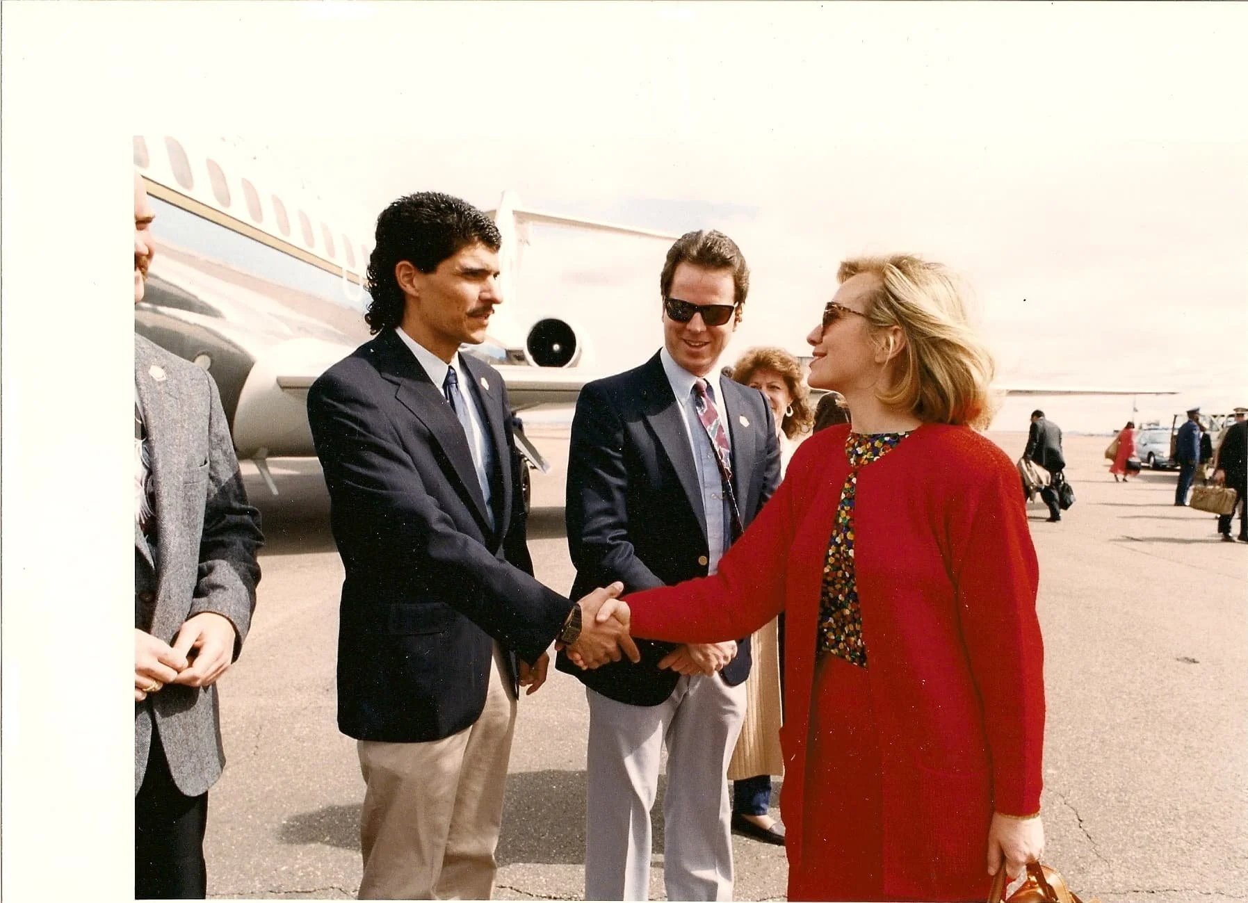 A young Bryan Lockerby shaking hands with Hillary Clinton outdoors near an airplane tarmac.