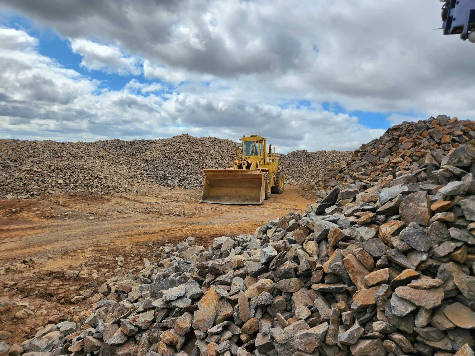 A yellow bulldozer on a dirt construction site with large piles of rocks and cloudy sky in the background.