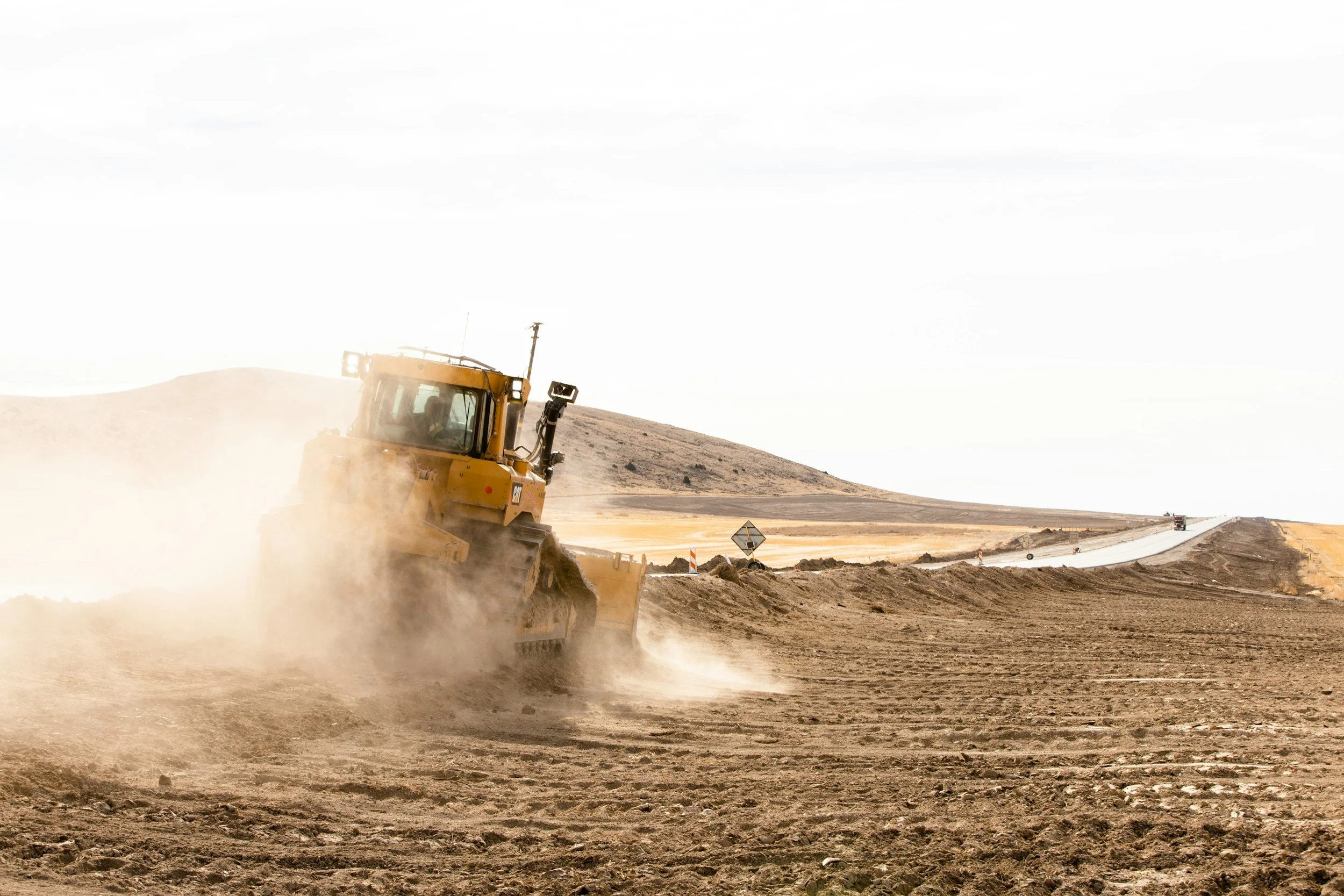 A yellow bulldozer working on a dirt construction site, kicking up dust as it moves soil along an open landscape with hills in the background.