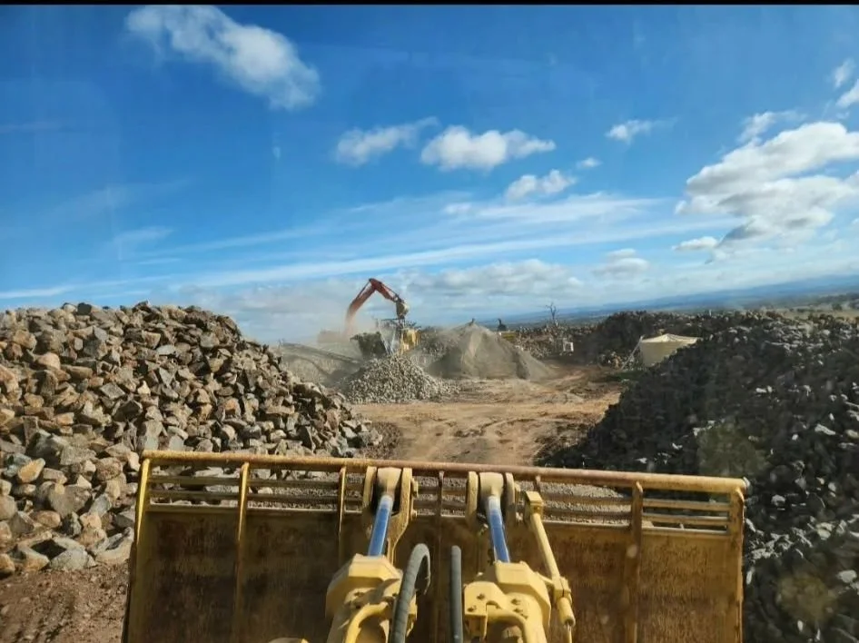 Construction site with a front loader moving rocks, an excavator in the background, piles of rocks on either side, under a partly cloudy sky.