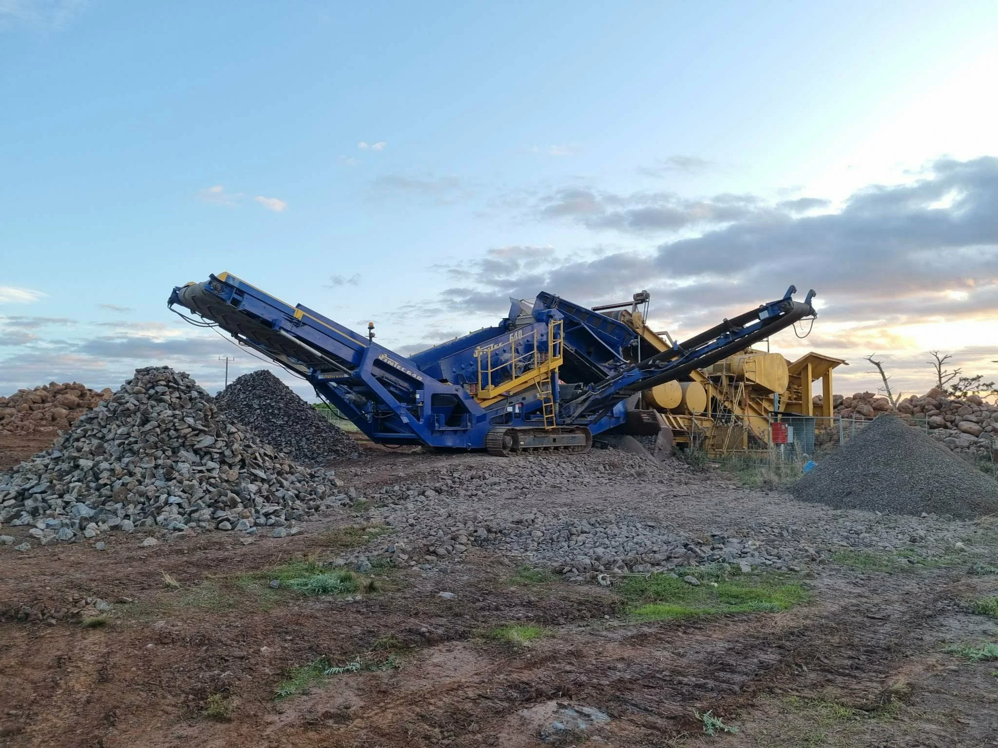 Large blue rock crushing machine operating outdoors at sunset with piles of rocks nearby.