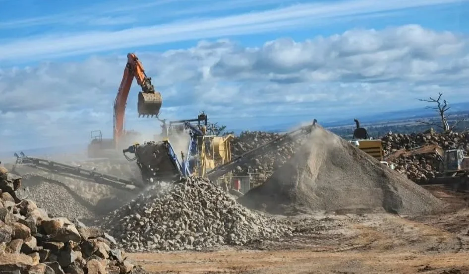 Construction site with an excavator moving rocks and gravel, dirt piles, and a blue sky with clouds in the background.
