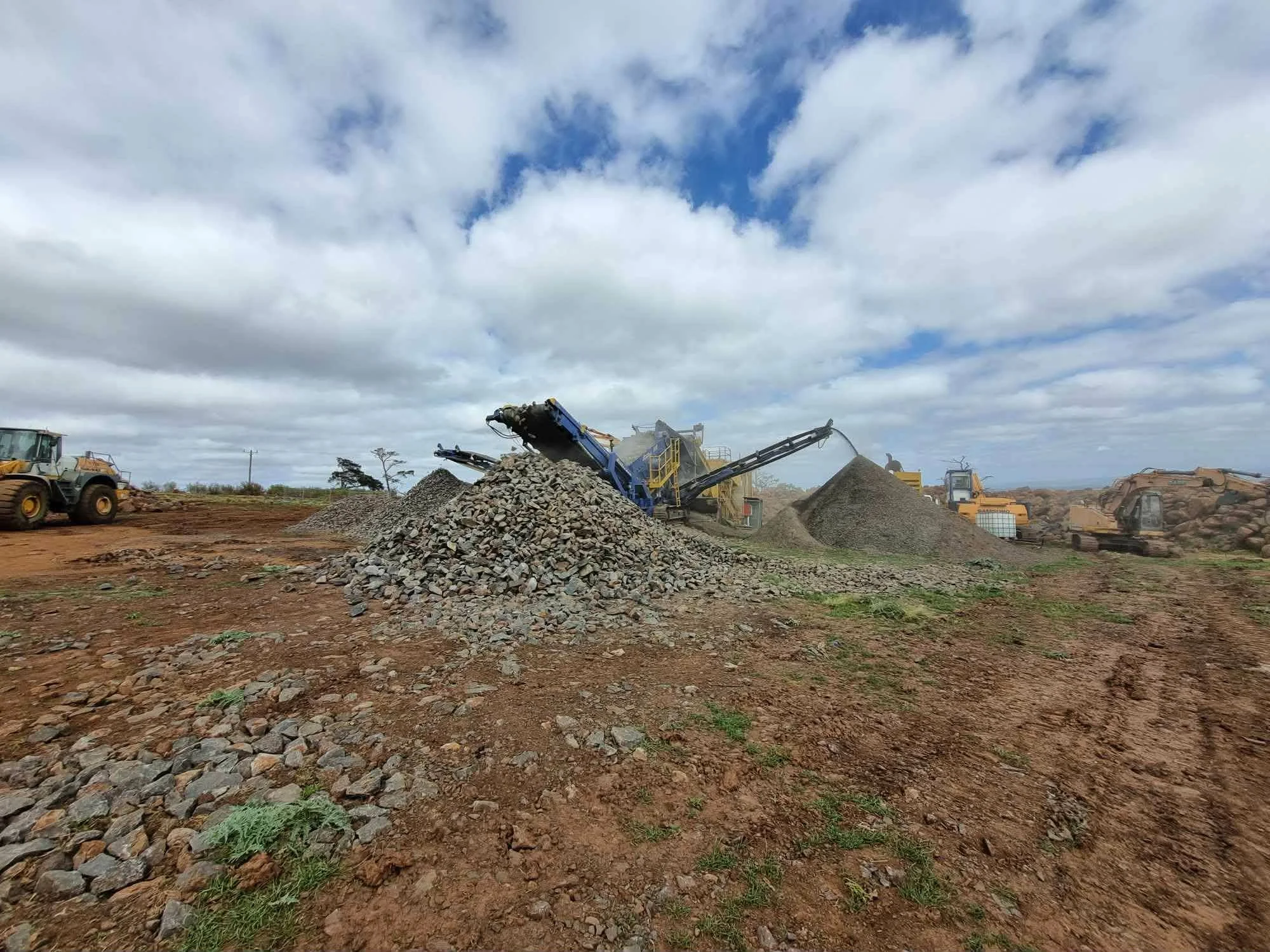 Construction site with industrial machinery sorting and piling rocks, with partly cloudy sky in the background, and some green vegetation on the reddish dirt ground.