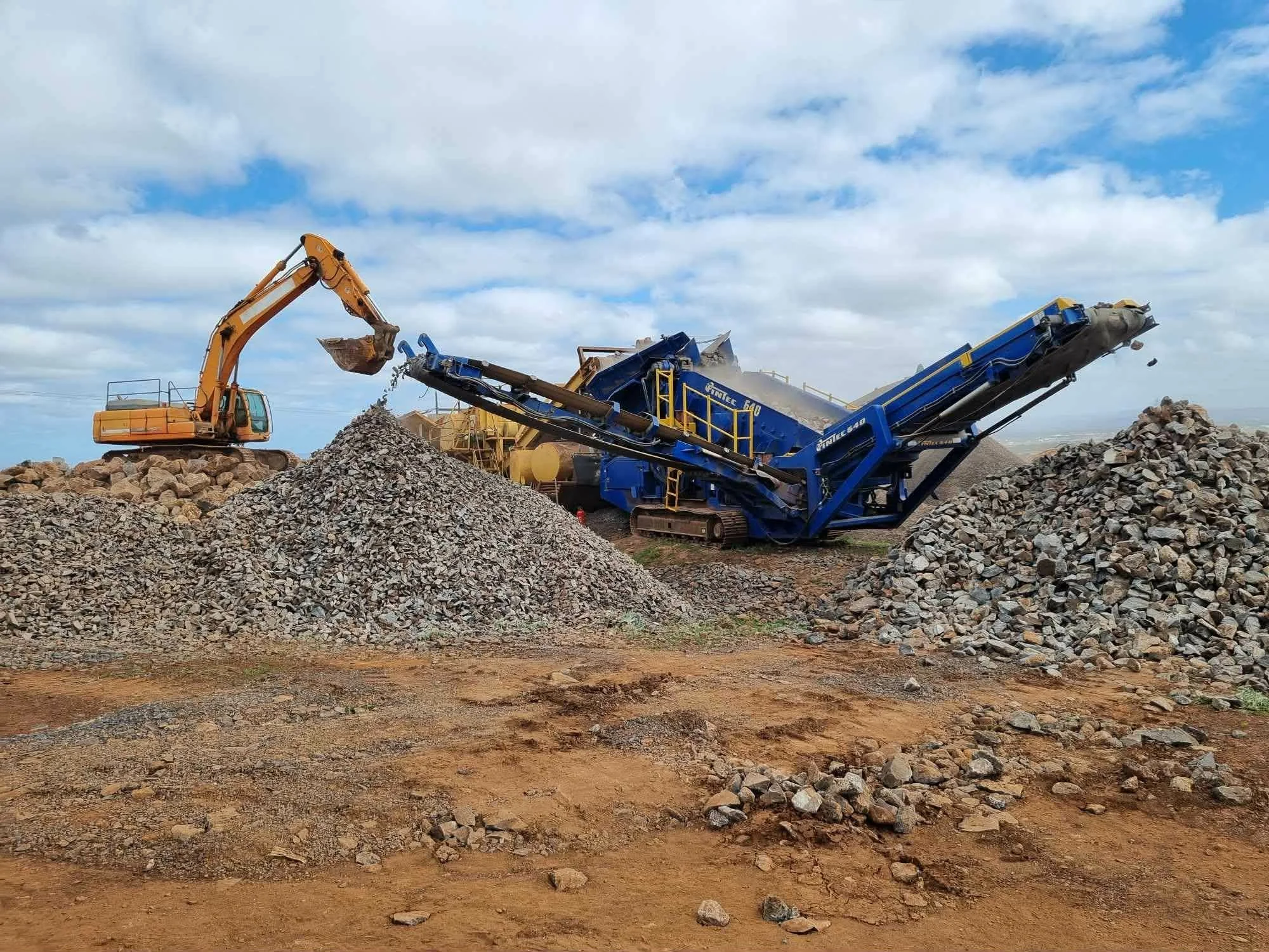 Construction site with an excavator loading rocks into a blue industrial rock crusher, dirt ground, cloudy sky