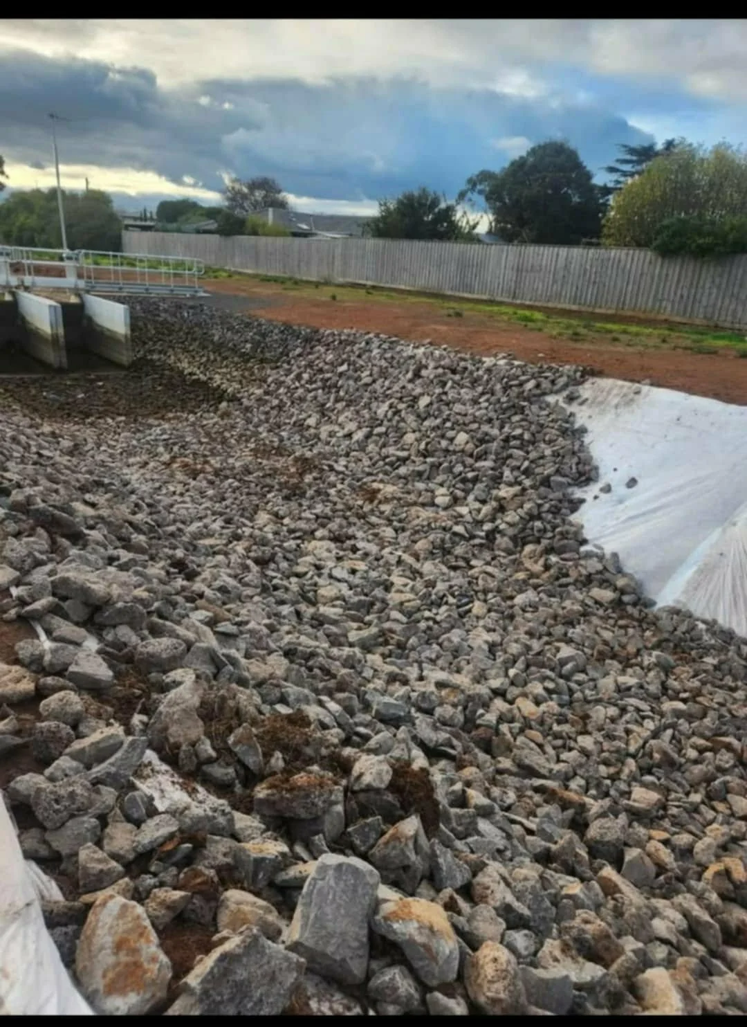 A construction site with rocks and gravel, a park gate, and a cloudy sky in the background.