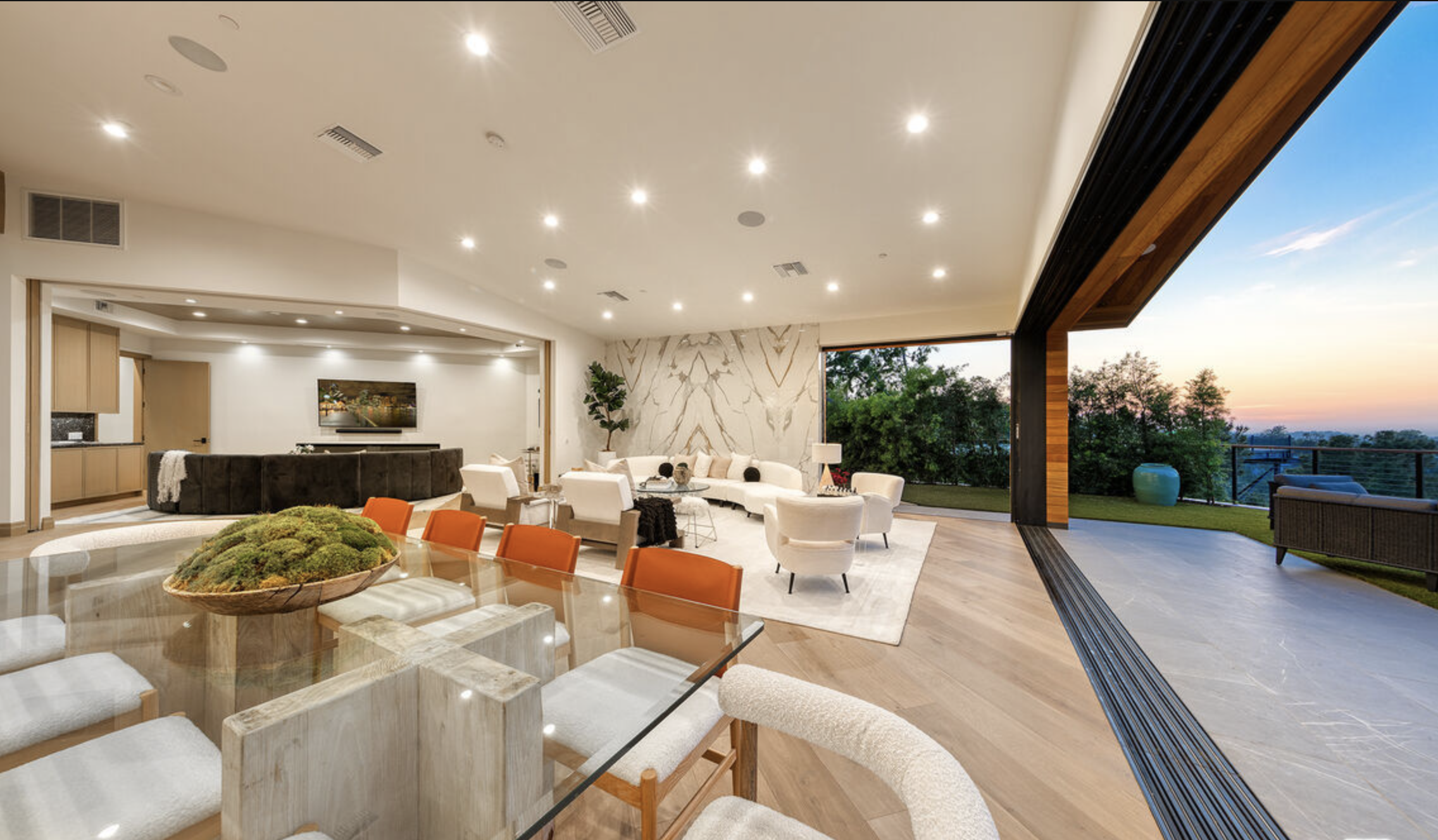 Modern living room with white furniture, a large glass dining table with orange chairs, and a view of the sunset outside on a patio