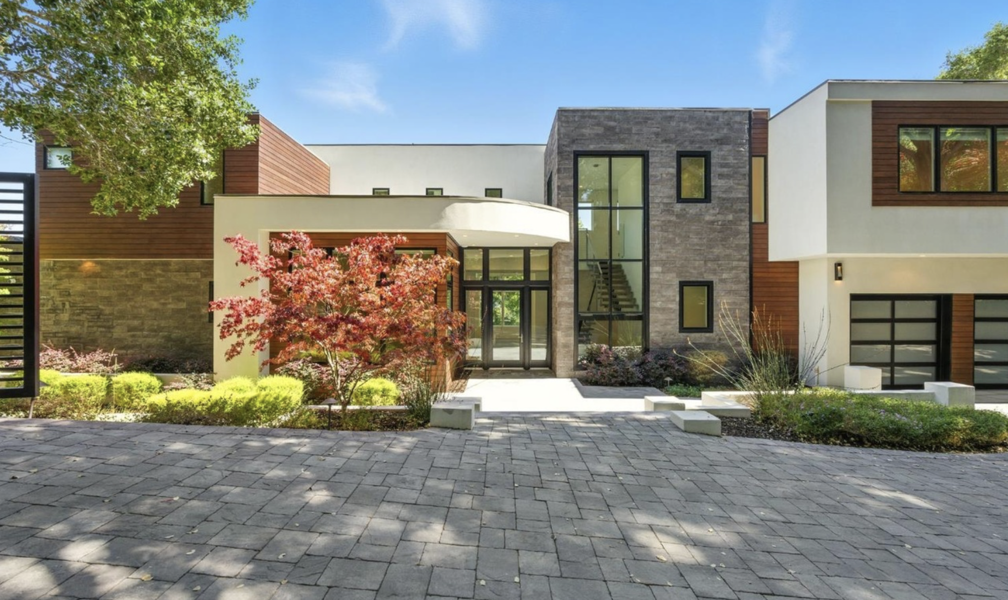 Modern house with a combination of wood, stone, and white exterior, surrounded by landscaping including a red-leafed tree and green bushes, under a clear blue sky.