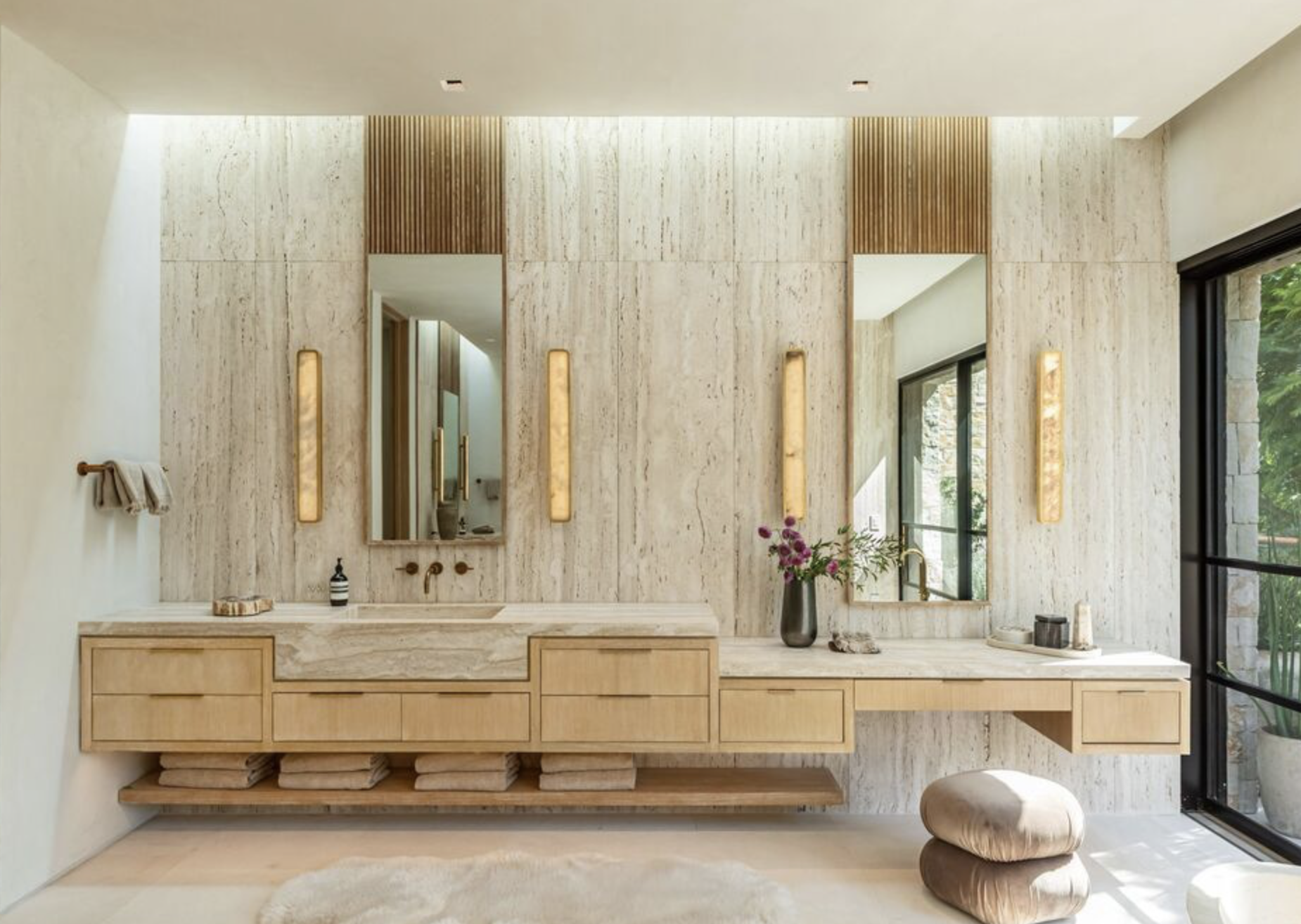 Modern bathroom with beige stone walls, wooden vanity with a marble countertop, two mirrors, and natural light from large windows.
