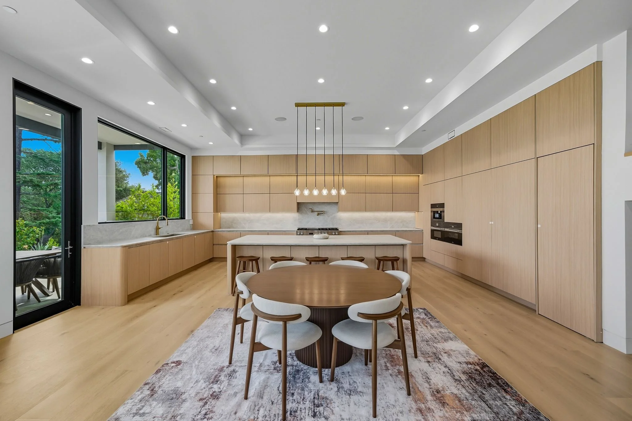 Modern kitchen with wooden cabinets, white marble backsplash, central island with bar stools, round dining table with white chairs, large window overlooking greenery, and hardwood floors.