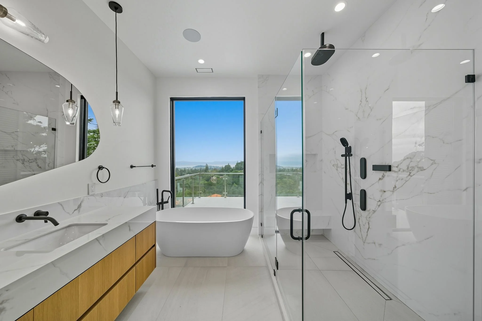 Modern bathroom with white marble walls and floor, a large glass shower with black fixtures, a freestanding bathtub, and a vanity with white countertop and wooden cabinets, large window with outdoor view.