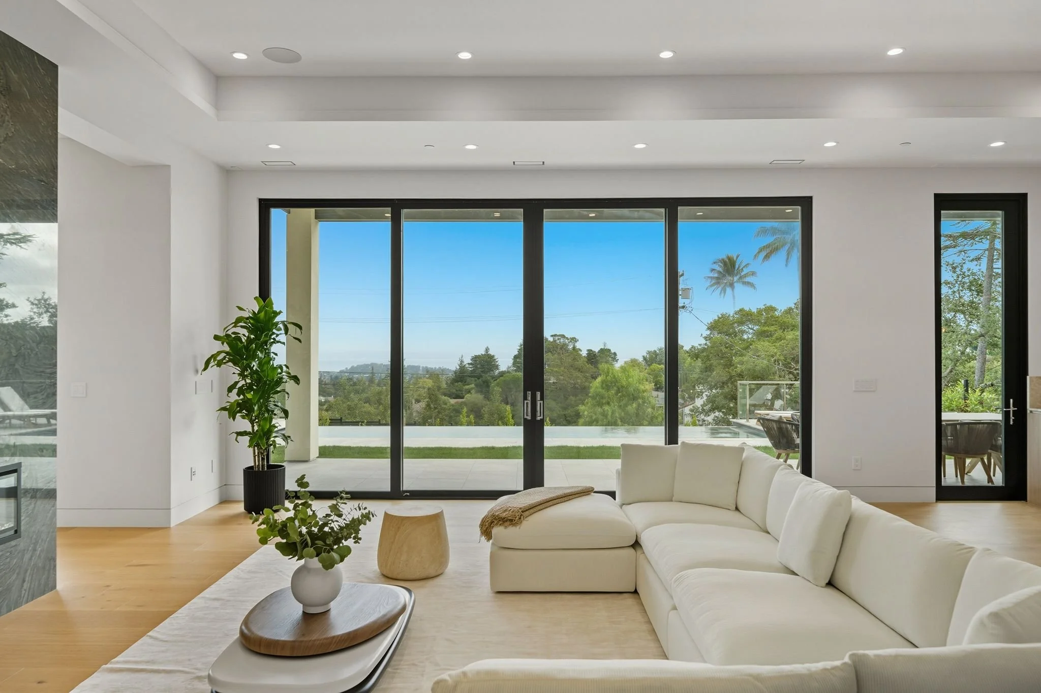 Modern living room with white sectional sofa, wooden coffee table, potted plant, and large sliding glass doors opening to a patio with a view of greenery and palm trees.