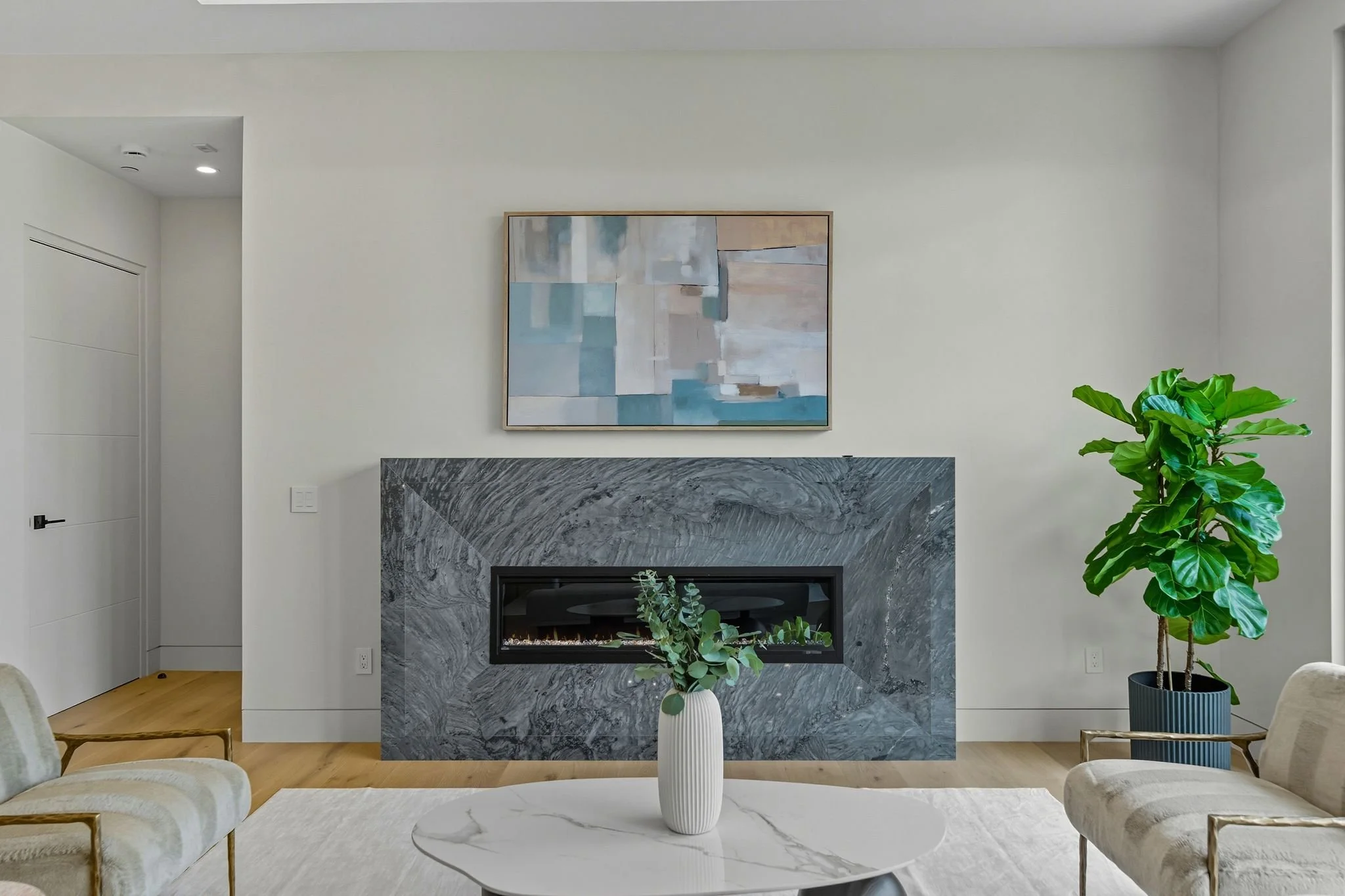 Modern living room with a fireplace framed in grey marble, a white vase with greenery on a white coffee table, and a large leafy plant in a dark blue pot to the right.