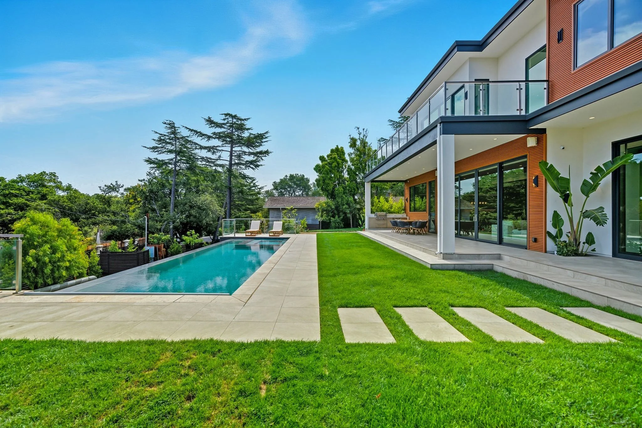 Modern house with large glass windows, outdoor patio, green lawn, and a swimming pool in a suburban neighborhood