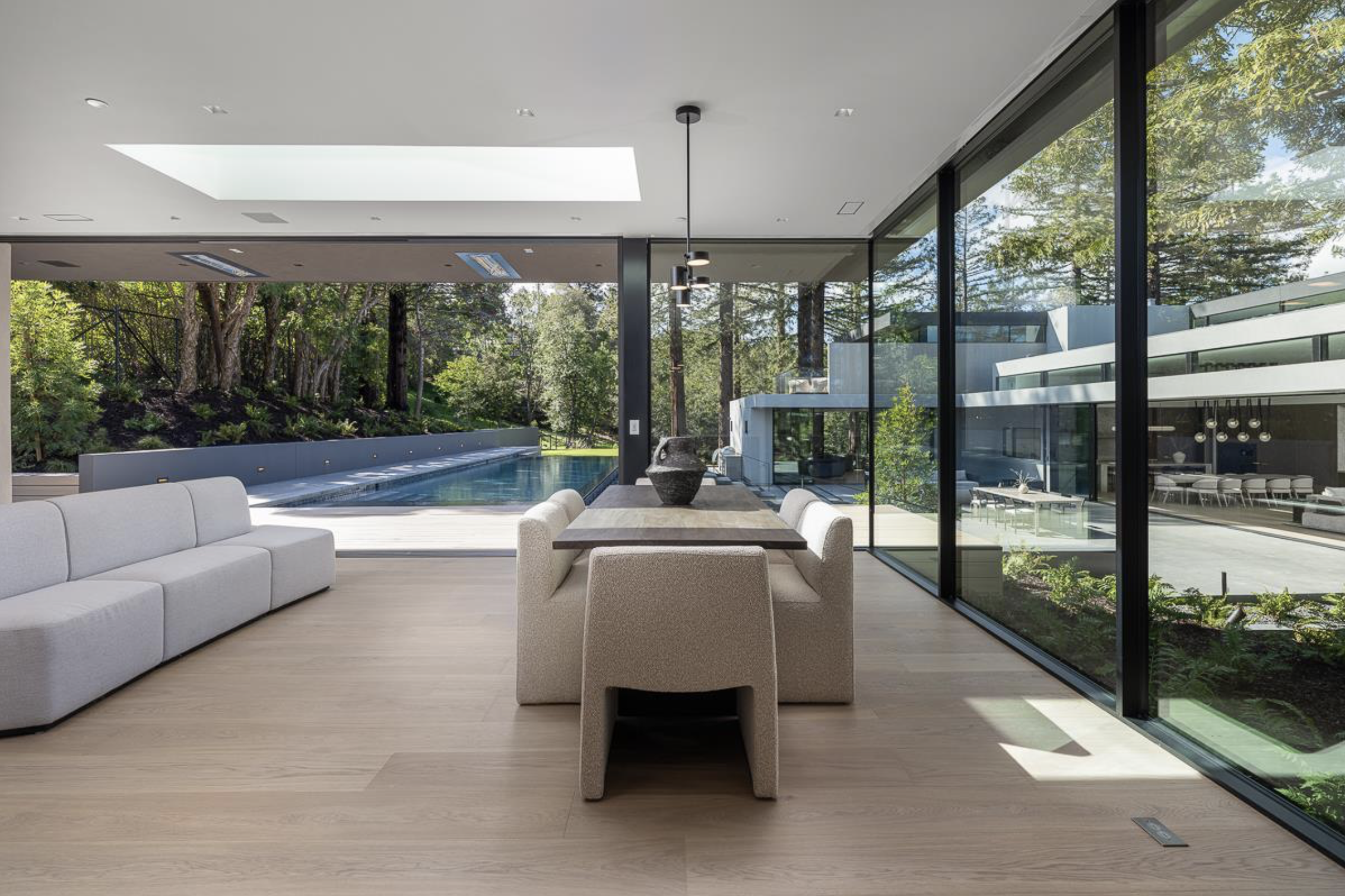 Modern living room with large glass windows, a dining table with six chairs, and a view of a pool and lush greenery outside.