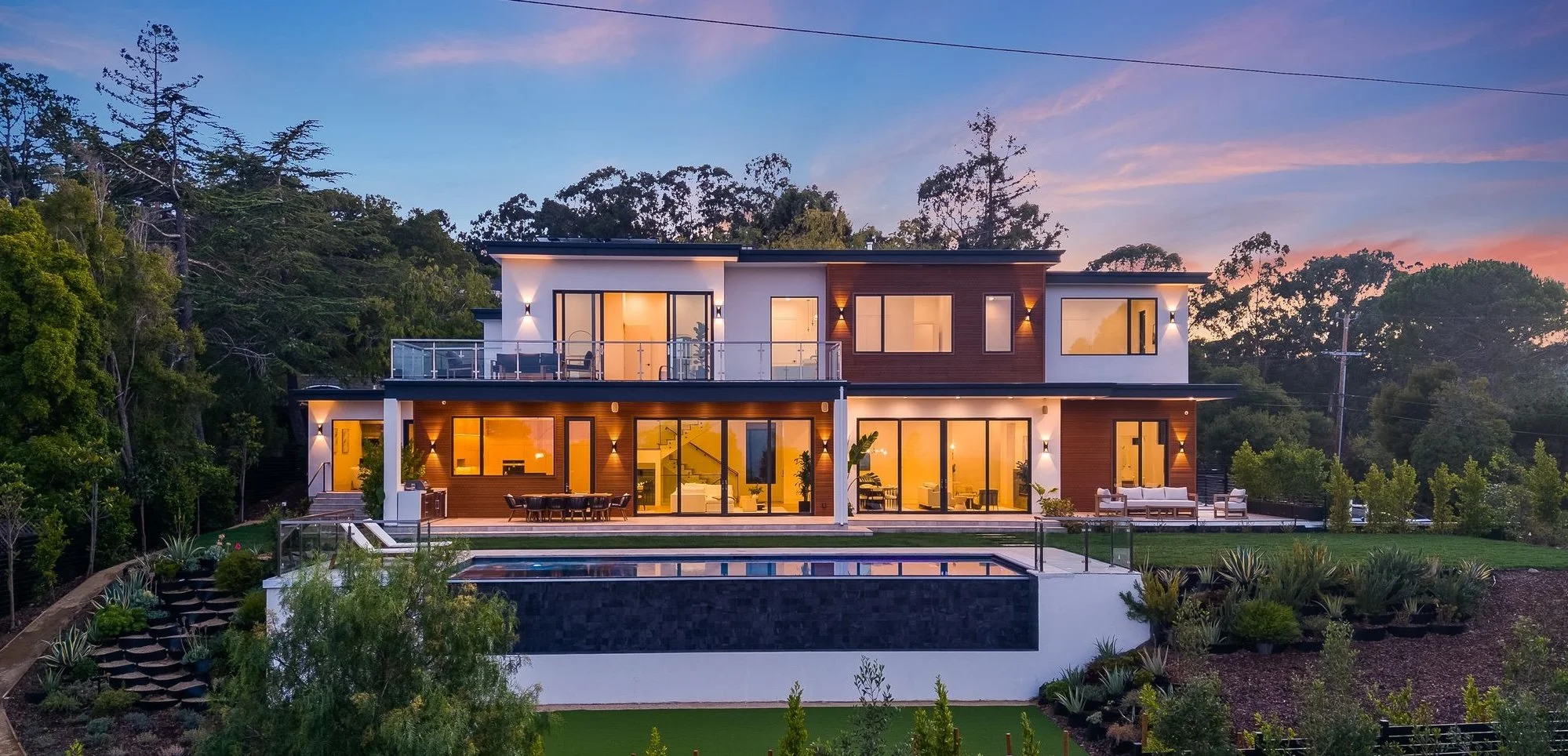 Modern two-story house with large glass windows and outdoor balcony, illuminated at dusk, surrounded by trees and landscaped yard with a pool.
