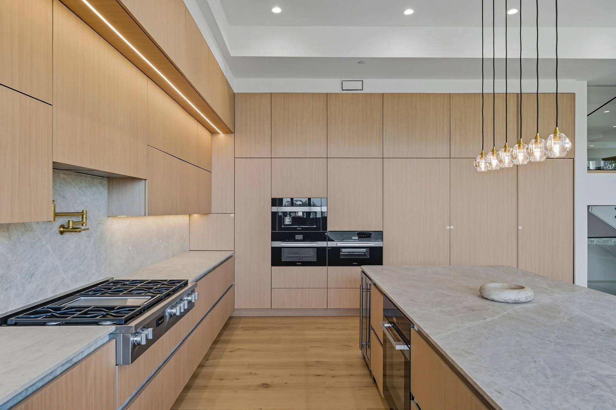 Modern kitchen with light wood cabinets, marble countertops, built-in oven and microwave, kitchen island, gold faucet, and a pendant light fixture.
