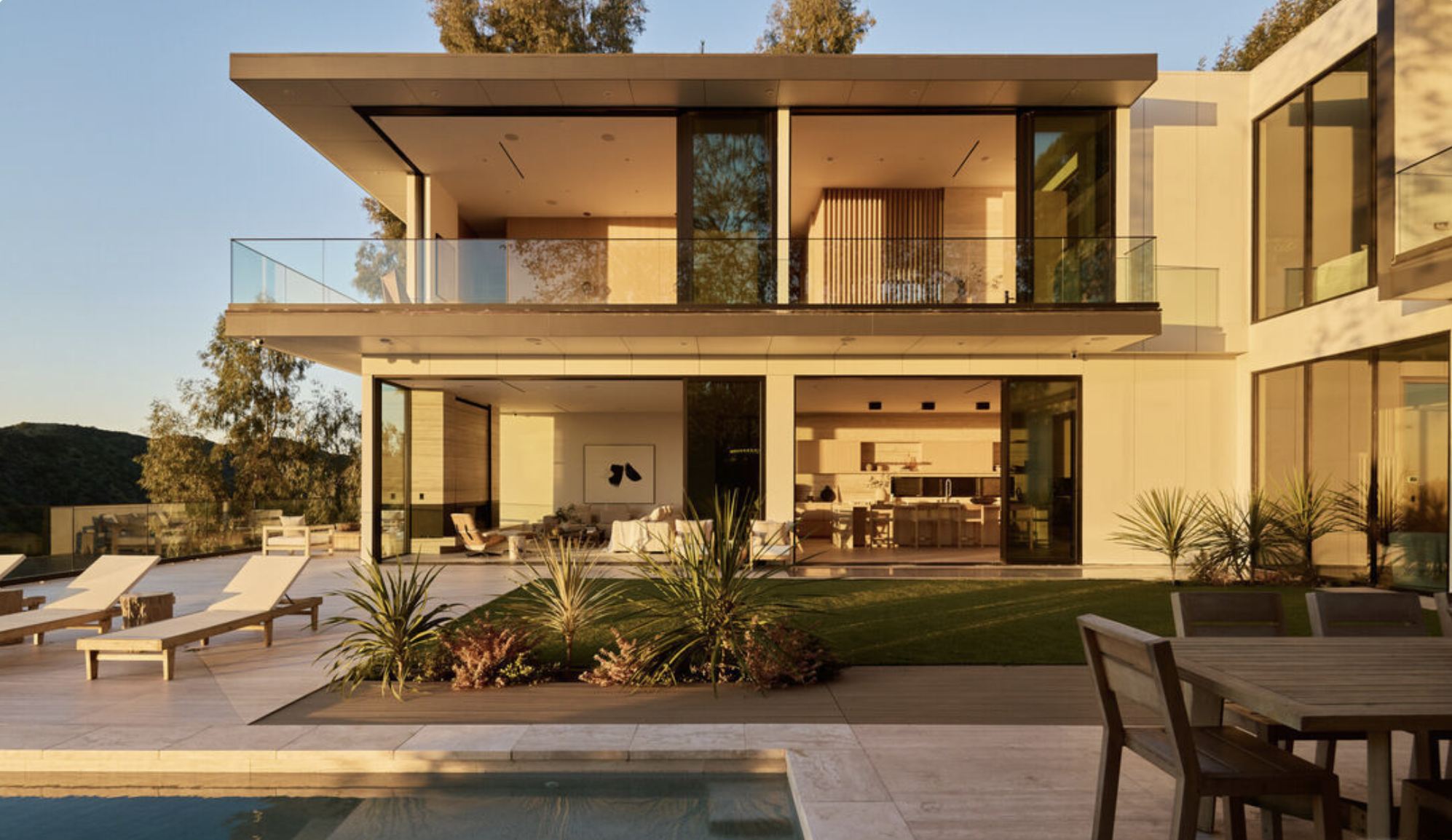Modern two-story house with large glass windows, outdoor wooden deck with lounge chairs, patio furniture, and a garden, during sunset.