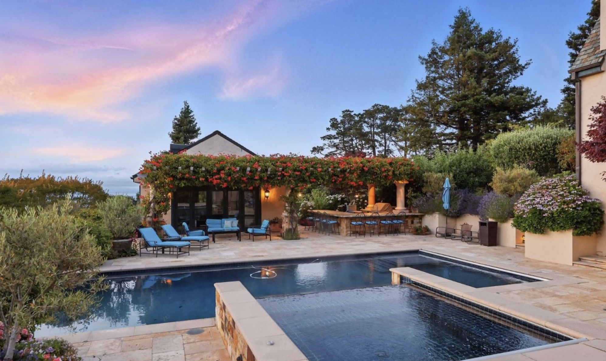 A backyard with a swimming pool, outdoor lounge chairs with blue cushions, a covered patio area with a bar and chairs, surrounded by flowering plants and trees, under a sky with pink and blue hues at sunset.