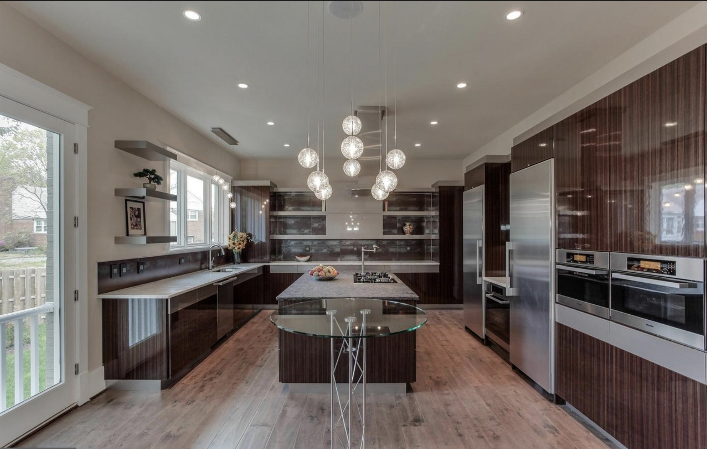 Modern kitchen with dark wood cabinets, stainless steel appliances, a marble island, and pendant lights hanging from a white ceiling. There is a glass table in front and windows that reveal a backyard.