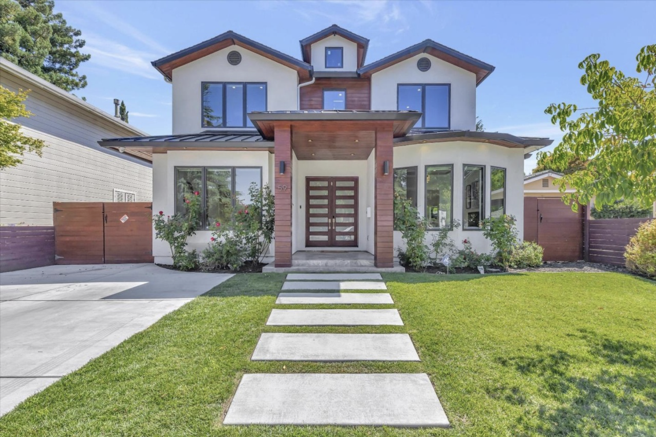Modern two-story house with white exterior walls, large windows, and a front porch with wooden accents. The house has a neatly landscaped front yard with a grass lawn and a stepping stone pathway leading to the front door. There's blooming shrubbery on either side of the entrance, and a wooden fence encloses the yard. Clear blue sky in the background.