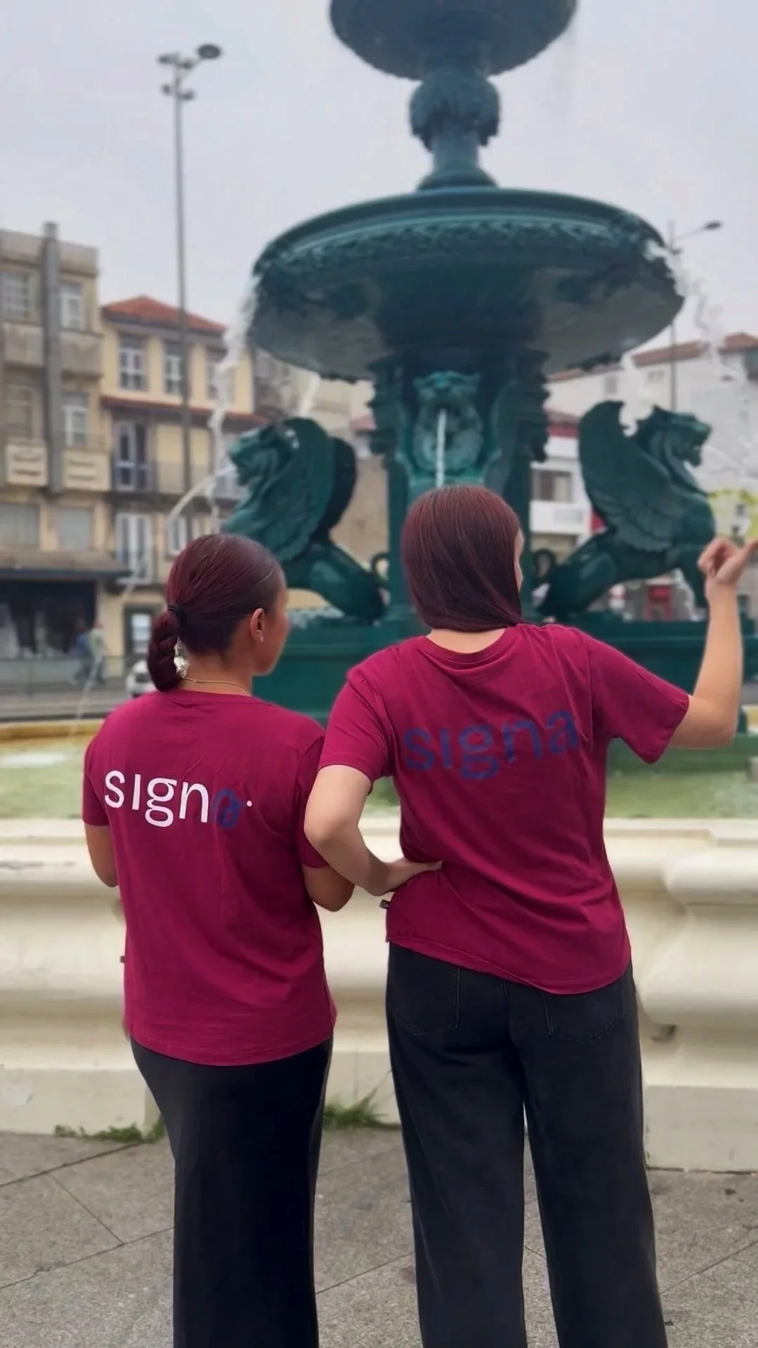 Duas mulheres de costas com t-shirts vermelhas com a palavra "SIGNA" nas costas, observando a fonte dos Leões no Porto.