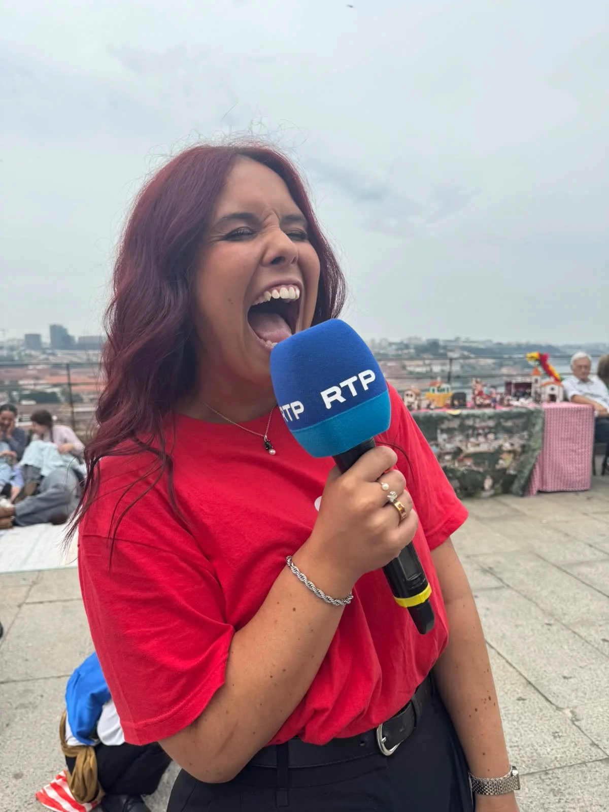 Criadora do Signa a sorrir e a segurar um microfone da RTP durante um evento ao ar livre, com pessoas ao fundo e vista de uma cidade ao longe.