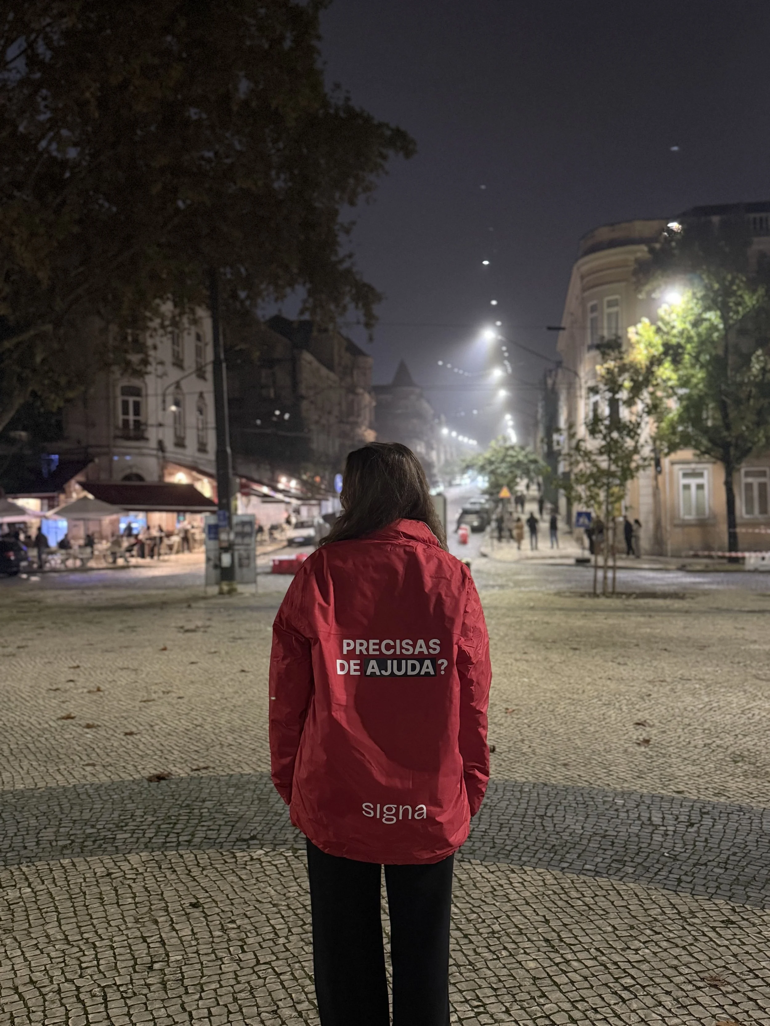 Pessoa parada de costas utilizando um casaco vermelho SIGNA com uma mensagem de apoio em português, "Precisas de Ajuda?", numa rua iluminada à noite.
