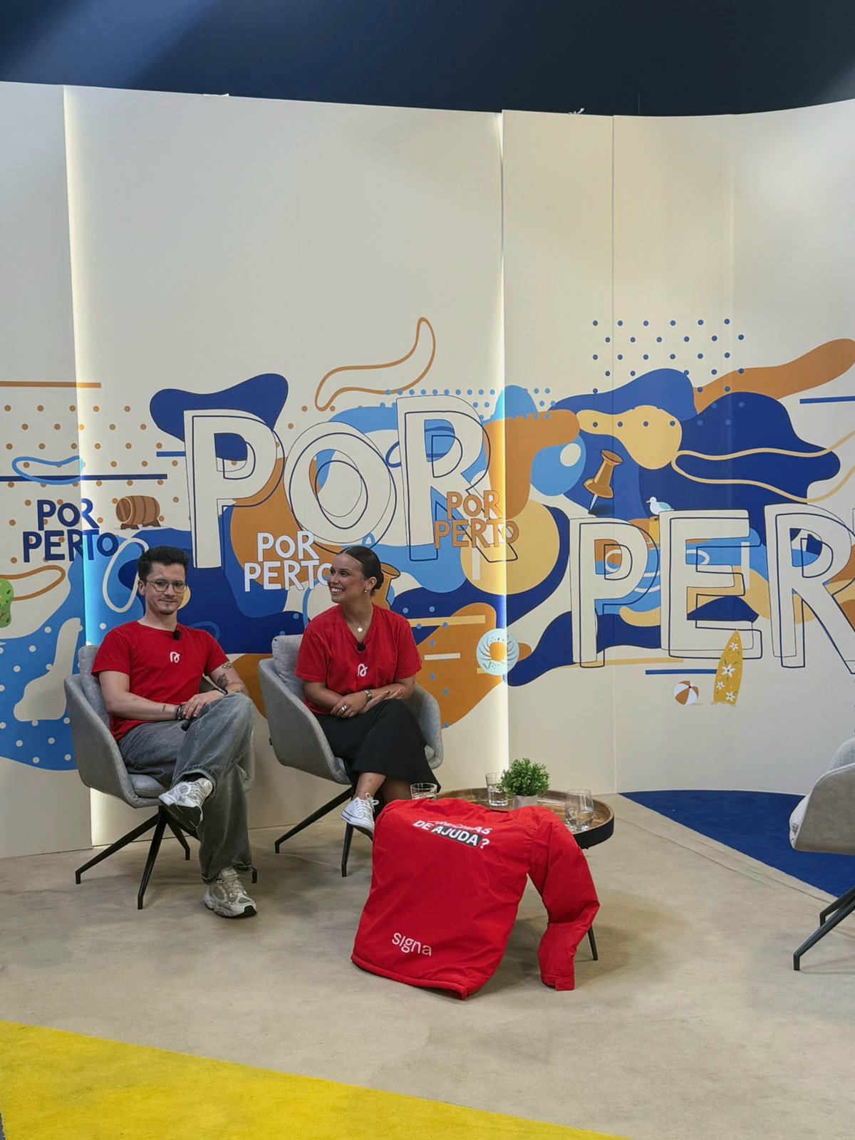 Dupla Signa sentada em cadeiras de frente a uma parede com arte e palavras "Por Perto", no programa do Porto Canal. Ambos usam camisetas vermelhas com o logo do Signa. 