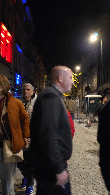 Pessoas a caminhar na rua à noite, com iluminação de lâmpadas de rua, enquanto está uma pessoa parada no centro utilizando um casaco vermelho SIGNA com uma mensagem em português, "Precisas de Ajuda?".