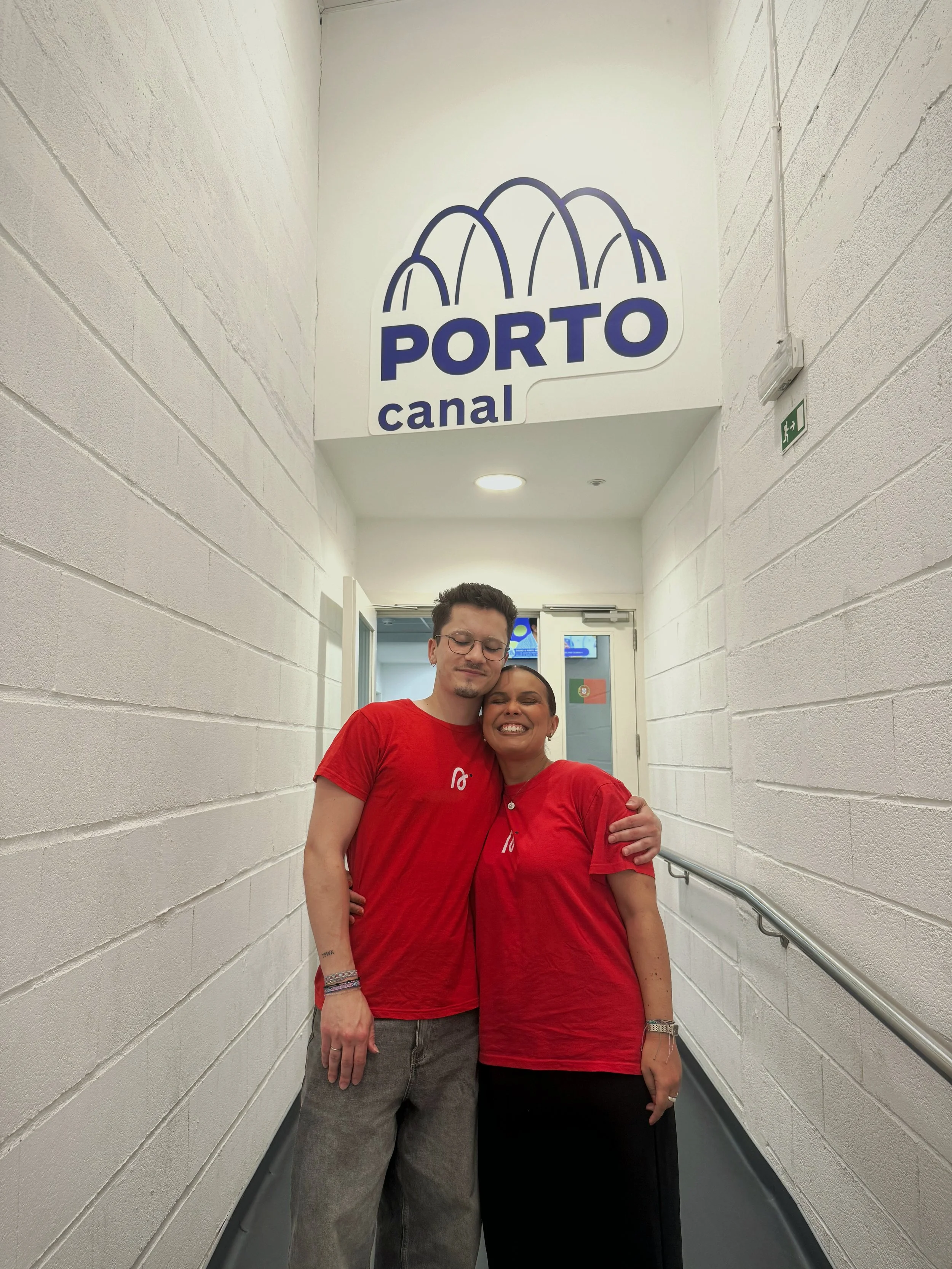 Dois indivíduos abraçados, ambos vestem t-shirts vermelhas com o logo Signa, na passagem de um corredor dos estúdios do Porto Canal.