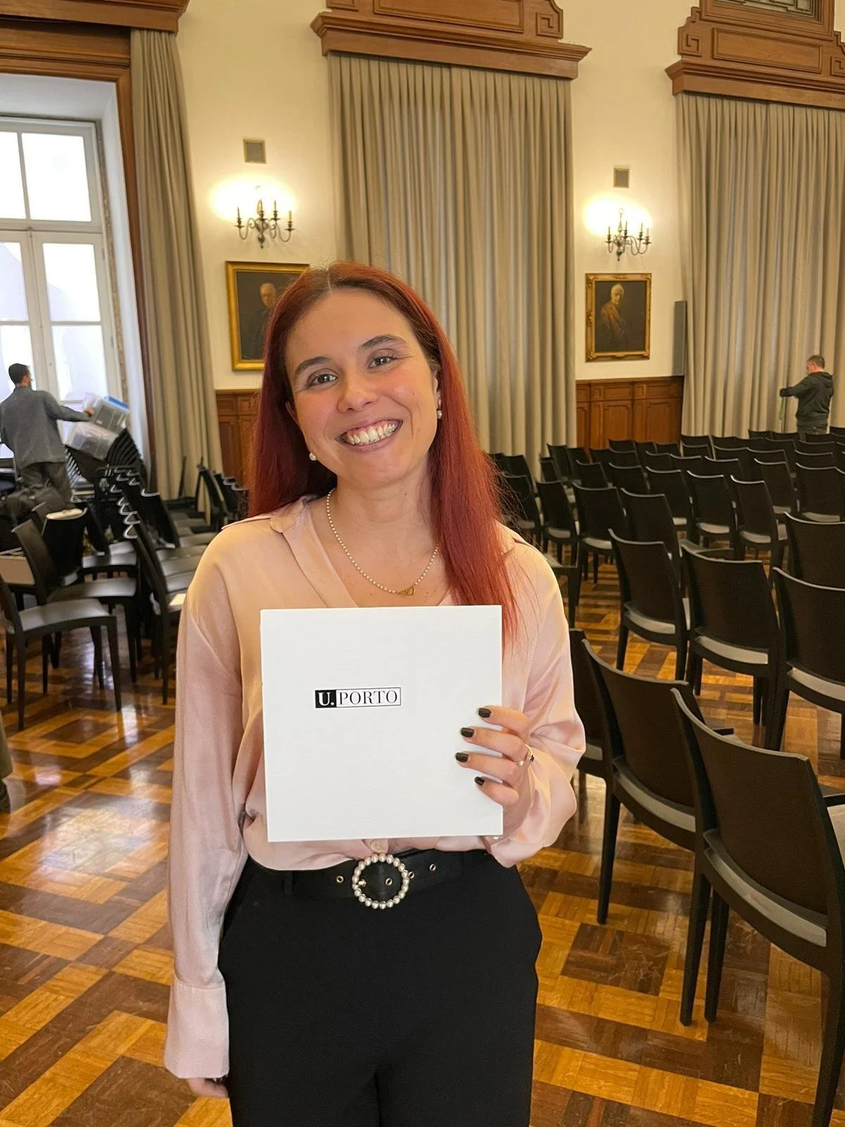 Mulher com cabelo vermelho, a CEO do SIGNA, segurando um envelope da U.PORTO numa sala de conferências da Reitoria da Universidade do Porto.. Ela sorri e usa roupa clara.