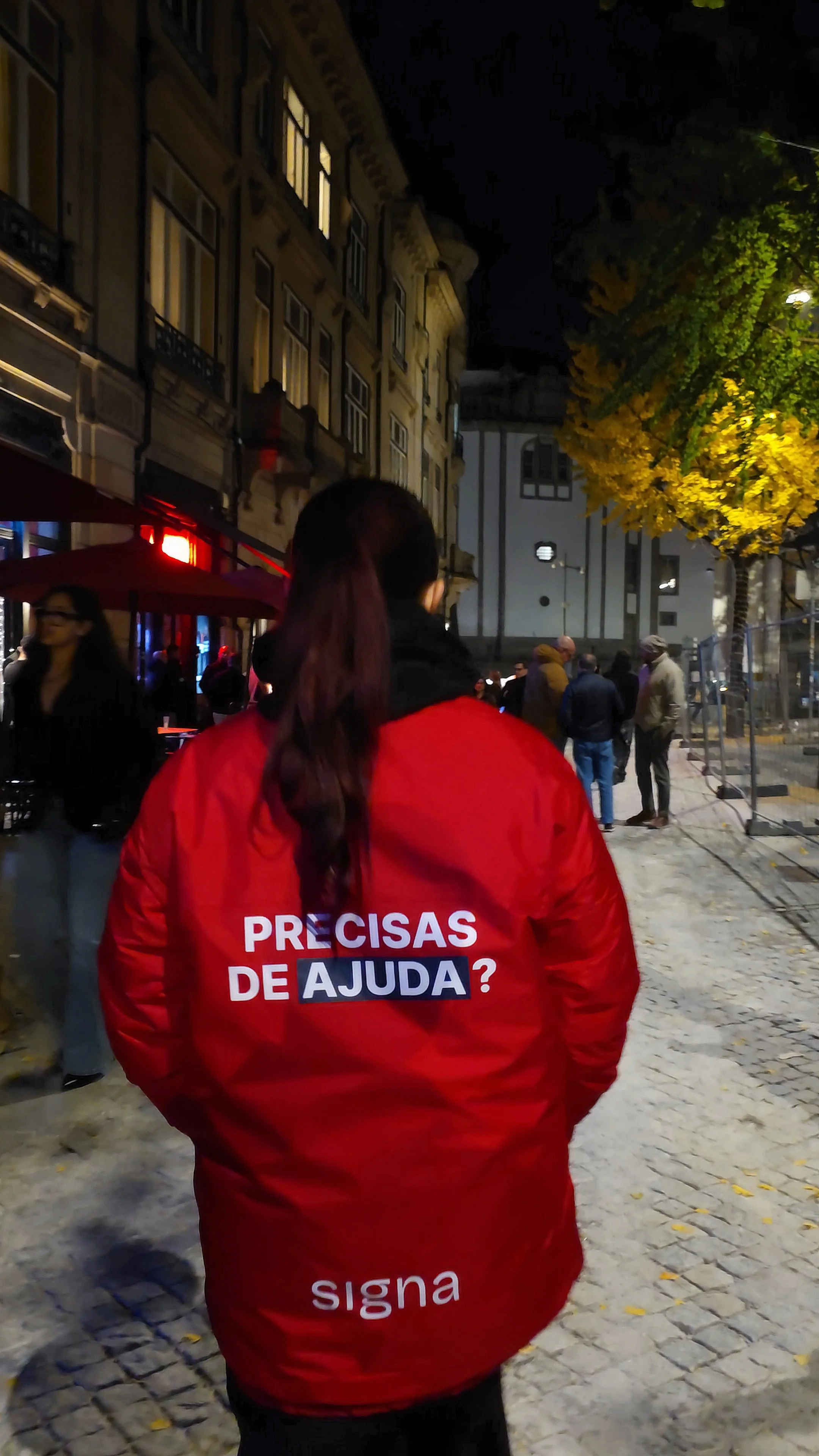 Pessoa de costas usando um casaco vermelho SIGNA com uma mensagem em português, "Precisas de Ajuda?", numa rua com outras pessoas, árvores com folhas amarelas e edifícios antigos ao fundo.