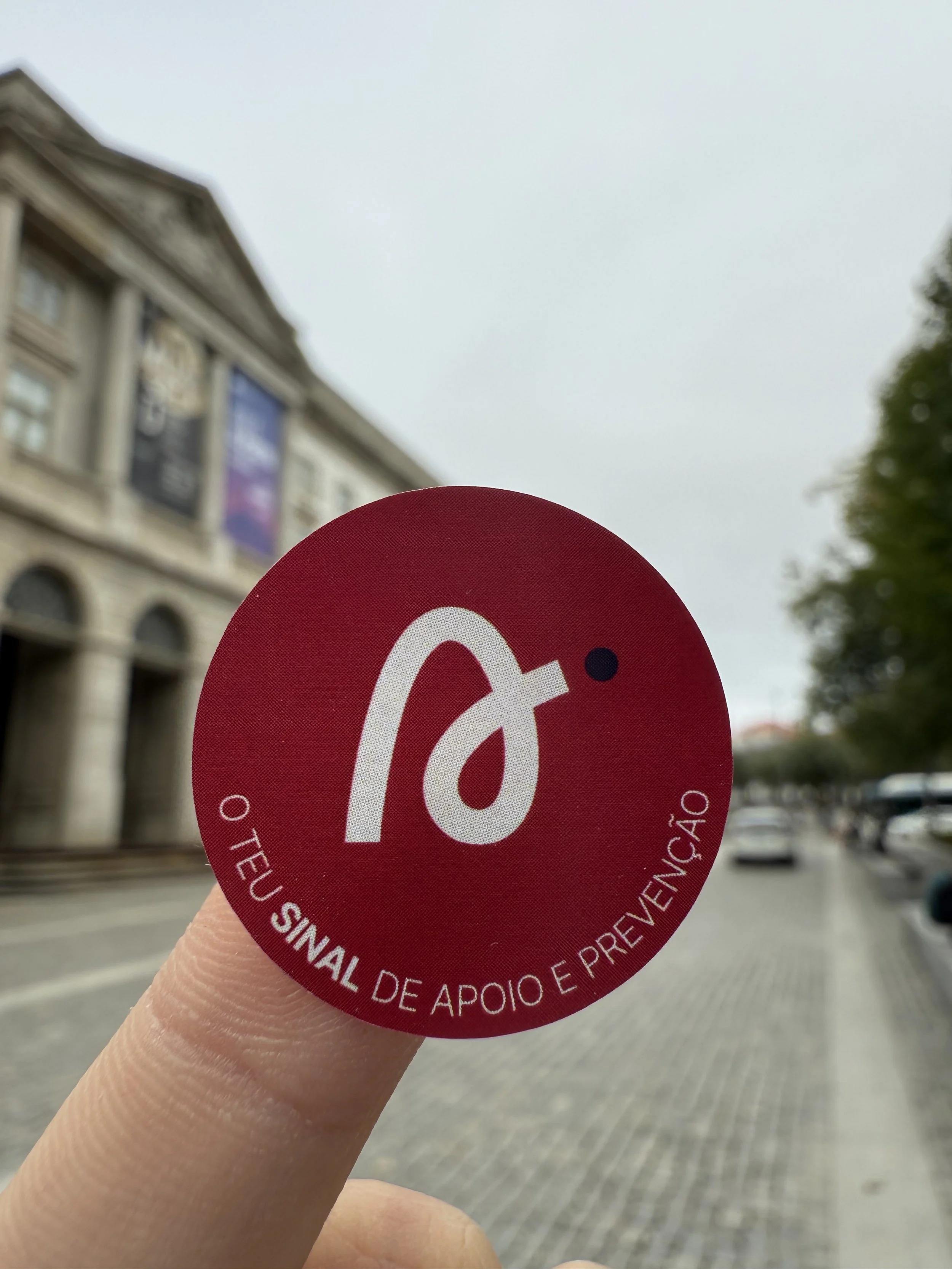Pessoa segurando um sticker vermelho do SIGNA com o símbolo de apoio e prevenção, na rua, com prédio e árvores ao fundo.