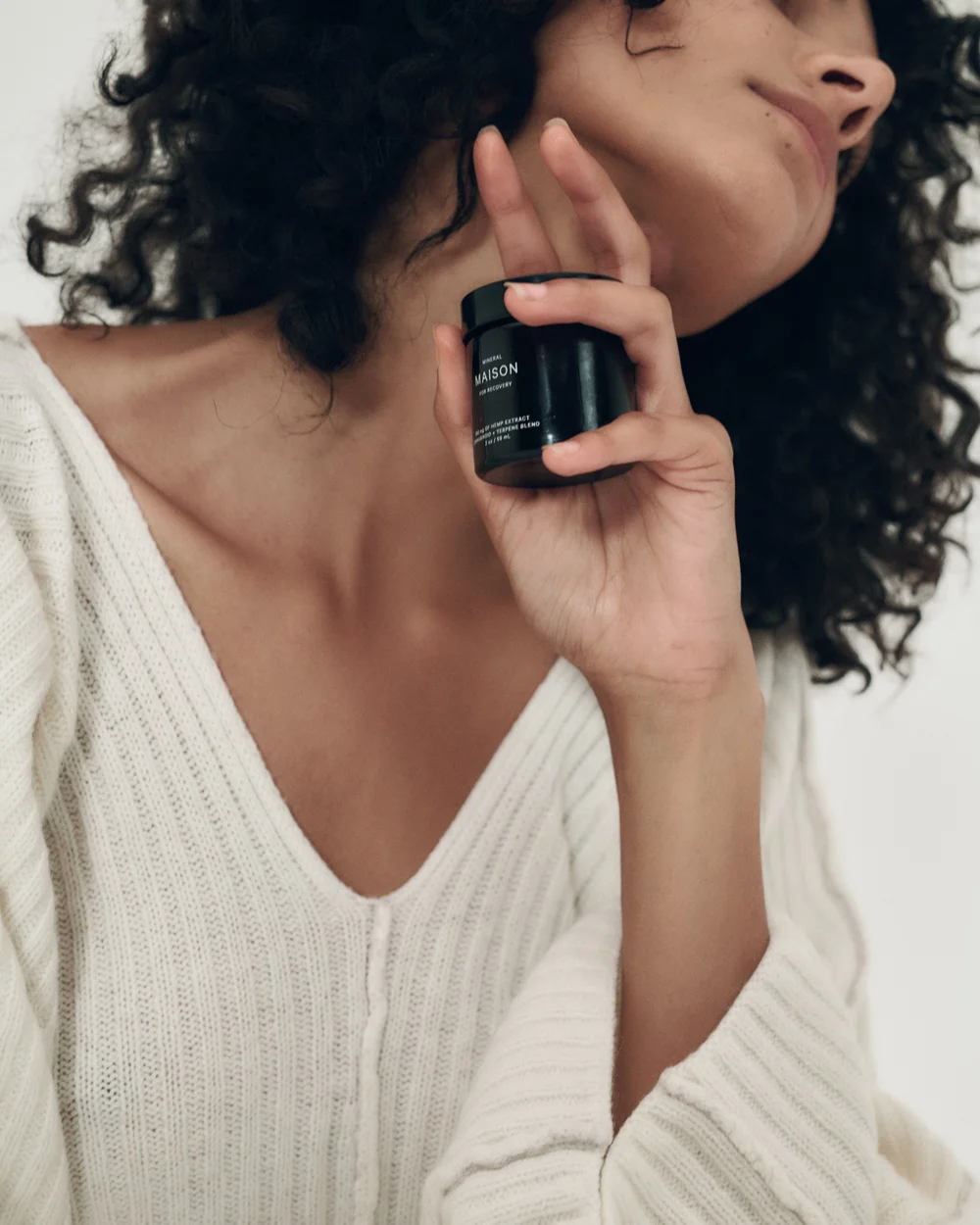 Close-up of a woman with curly hair holding a small black jar of skincare product near her face, wearing a cream-colored knit sweater.