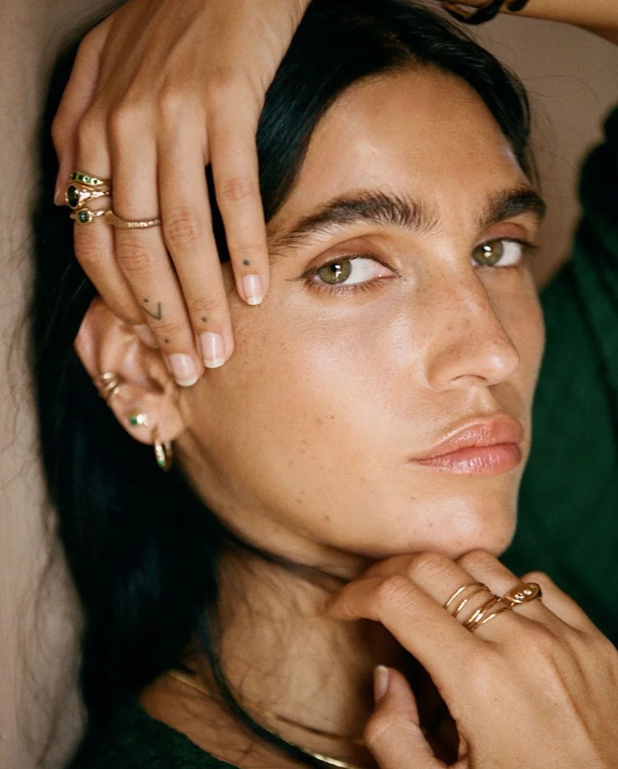 A woman with dark hair, multiple rings, and earrings, lying down with her hand on her face, wearing a green shirt.