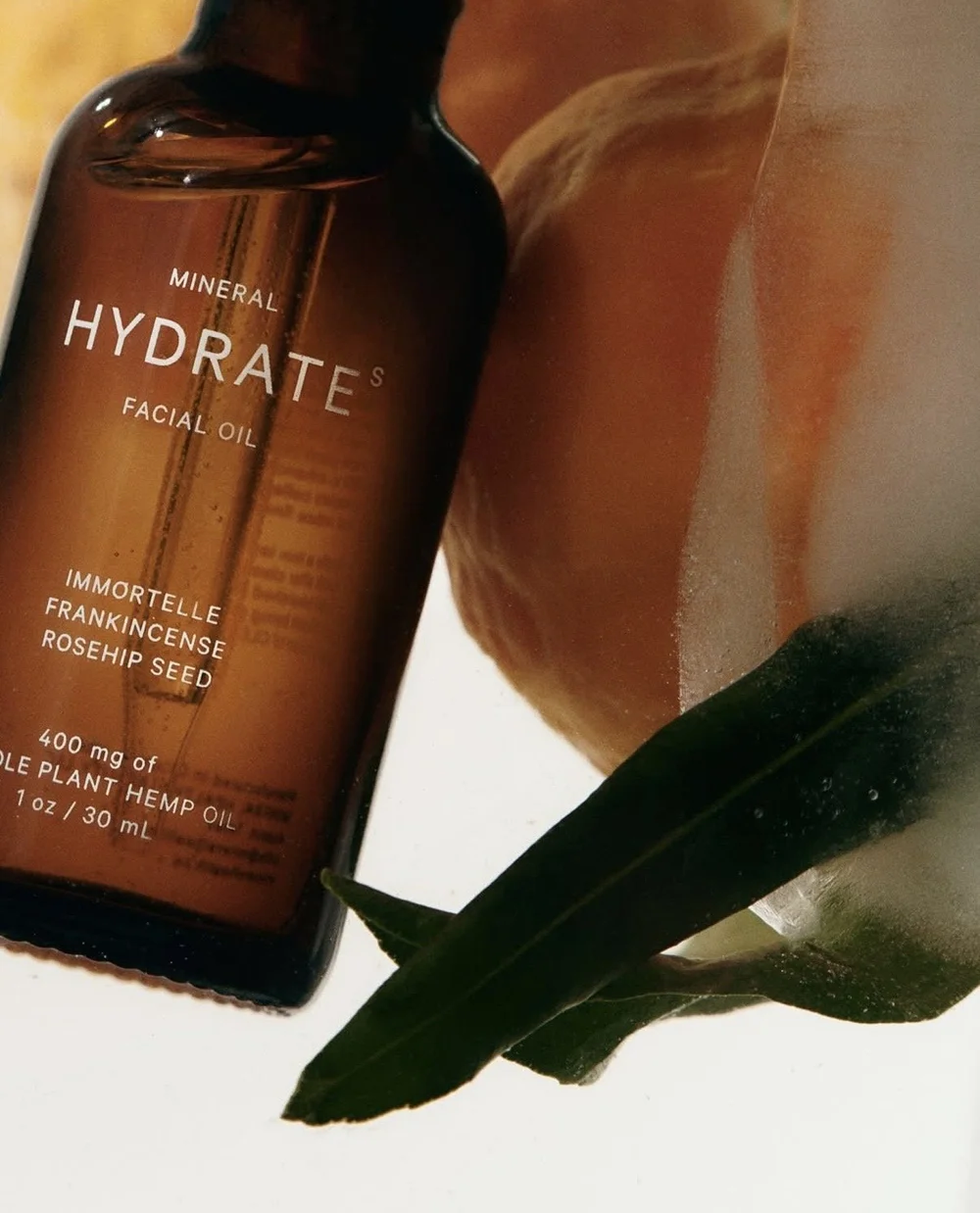 A bottle of mineral hydratés facial oil with a dark amber color, a green leaf, and a partially peeled orange.
