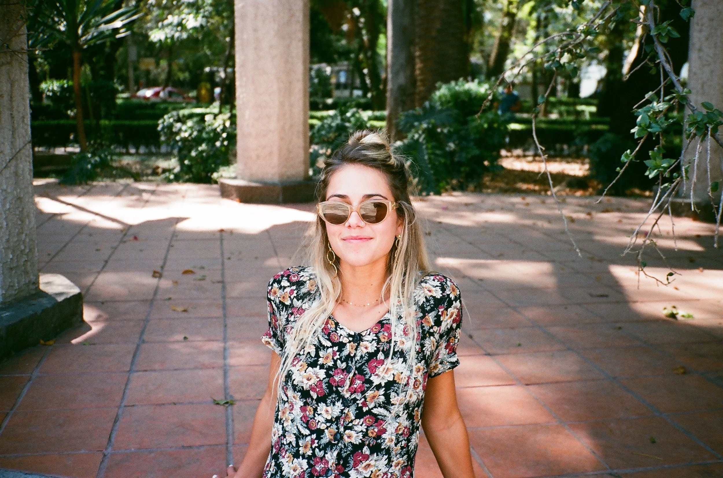 A young woman with long blonde hair wearing sunglasses and a floral dress stands outdoors, smiling gently at the camera, with trees and plants in the background.